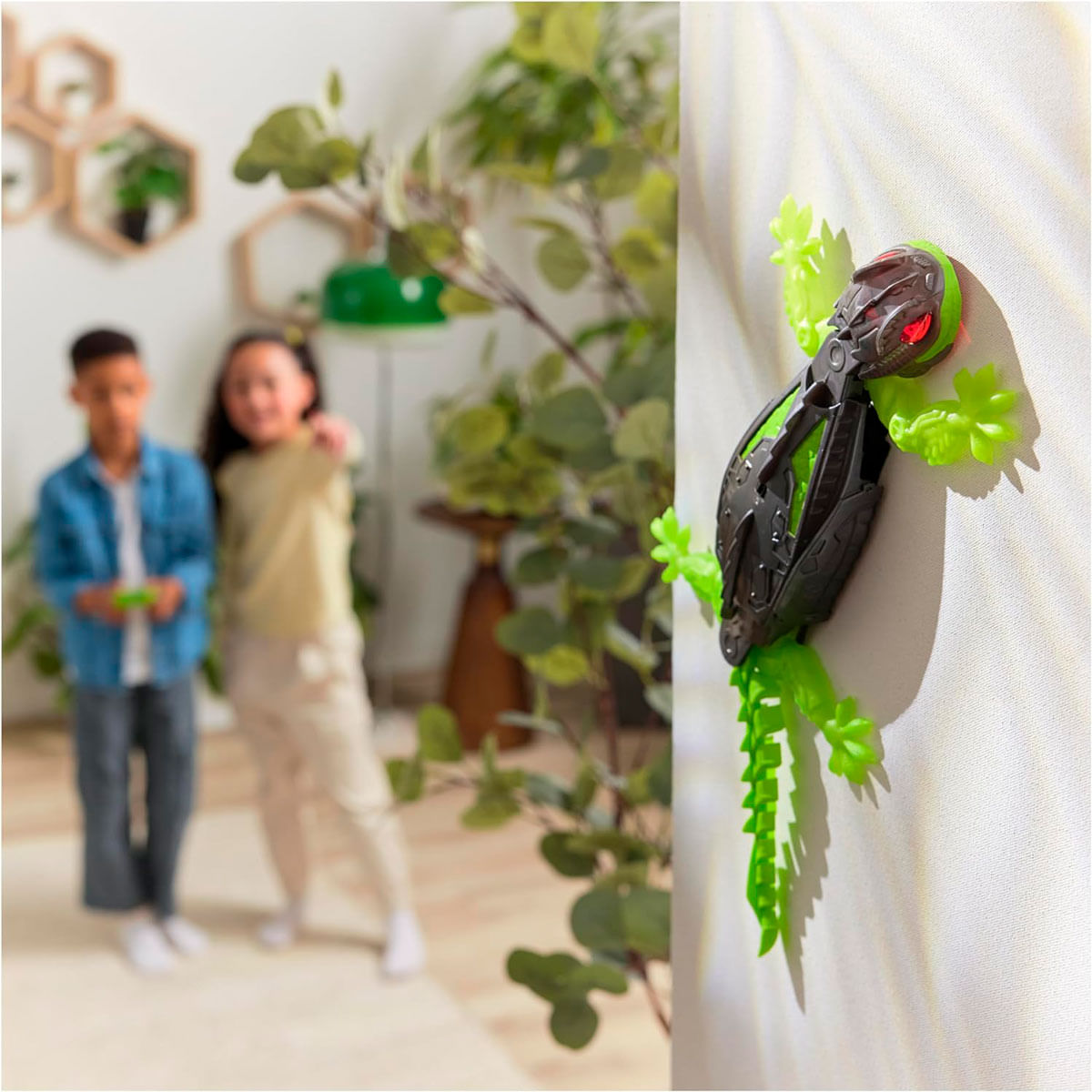 Un insecto de juguete está pegado a una pared. Dos niños están en el fondo borroso. Hay plantas verdes en el lado izquierdo.