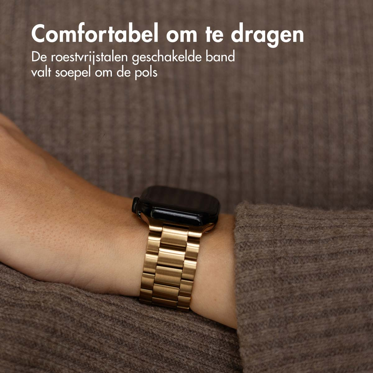 A person wearing a gold watch with a linked band. The watch is on the wrist, with a brown sweater in the background.