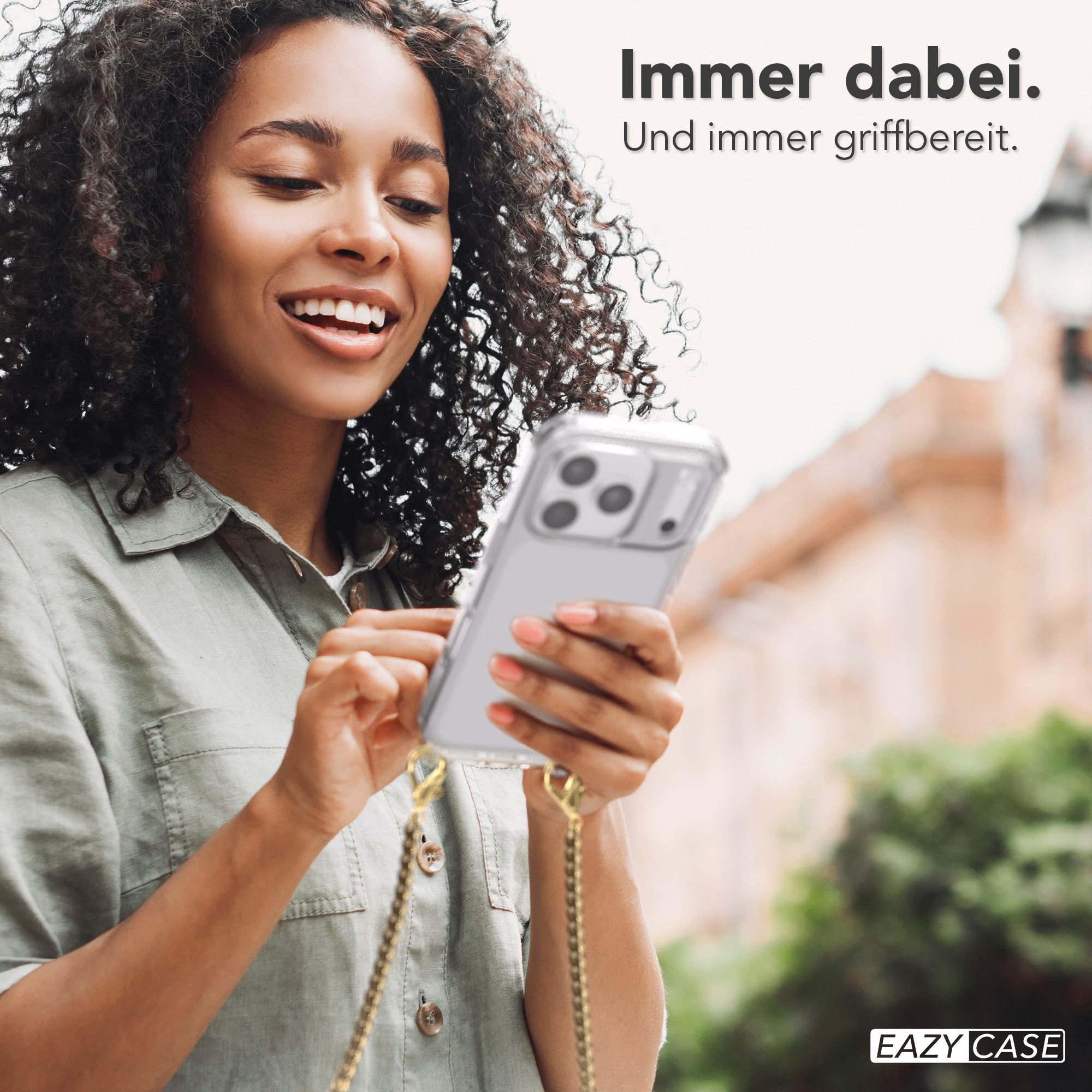 Eine lächelnde Frau hält ein Telefon mit einer transparenten Hülle. Goldene Kette befestigt. Hintergrund ist unscharf.