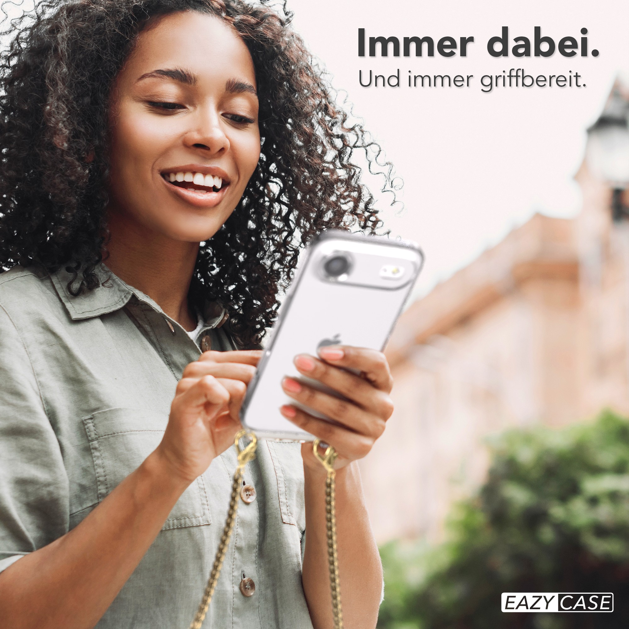 Lächelnde Frau hält ein Telefon in einer transparenten Hülle. Goldene Kette, grünes Hemd. Hintergrund unscharf.