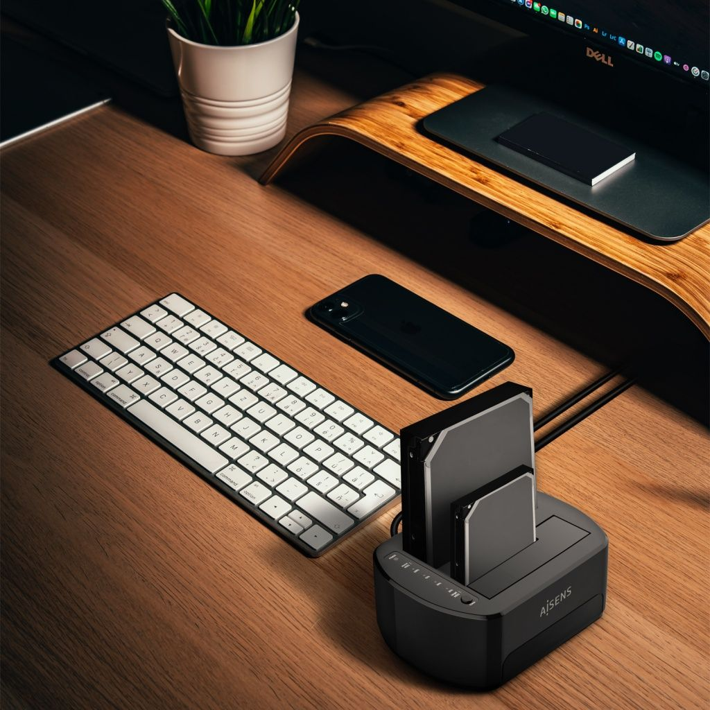 Un escritorio con teclado, teléfono y una base para disco duro. También se ve una planta en una maceta blanca.