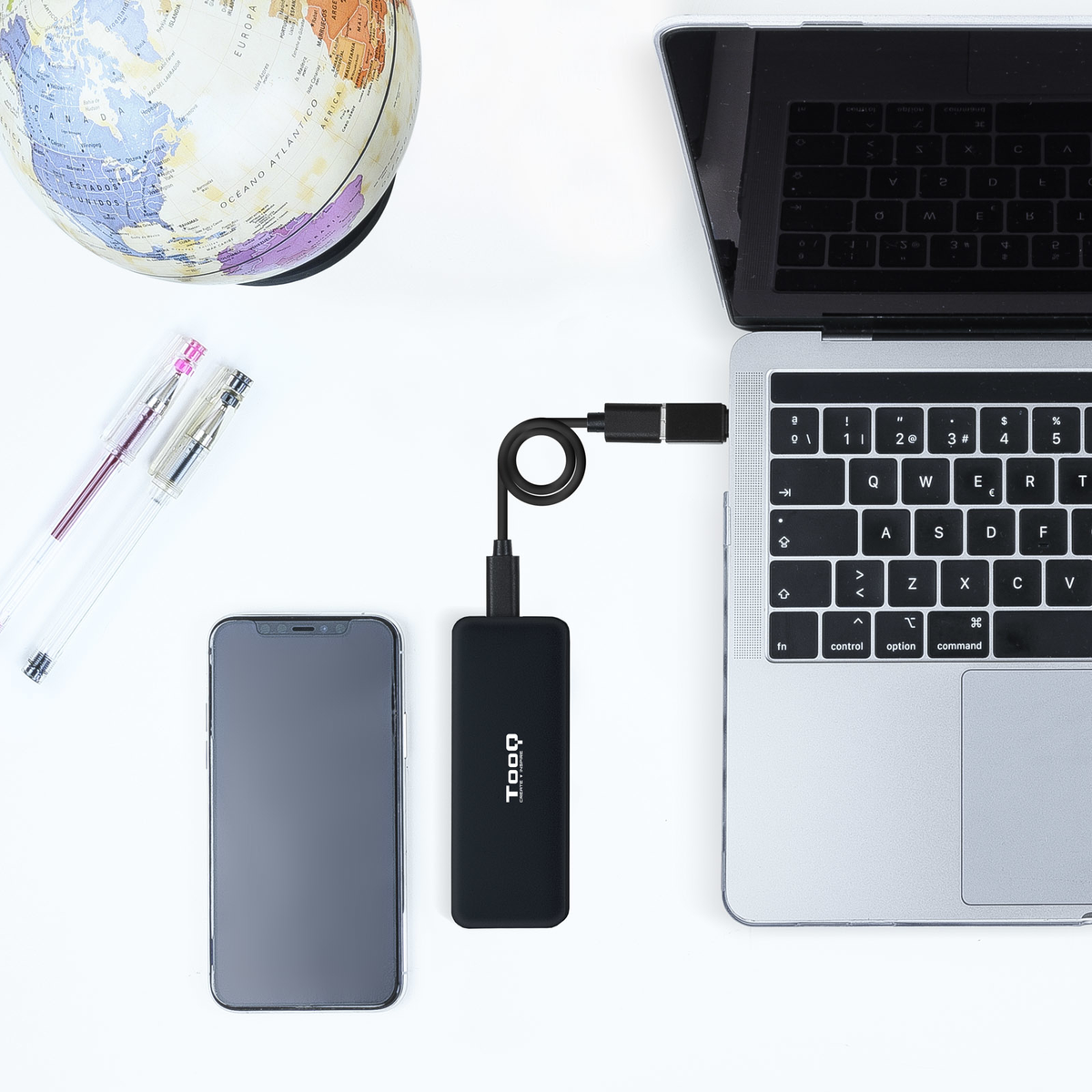 Un smartphone, un portátil, un globo terráqueo, bolígrafos y un banco de energía negro con cable están sobre una superficie blanca.