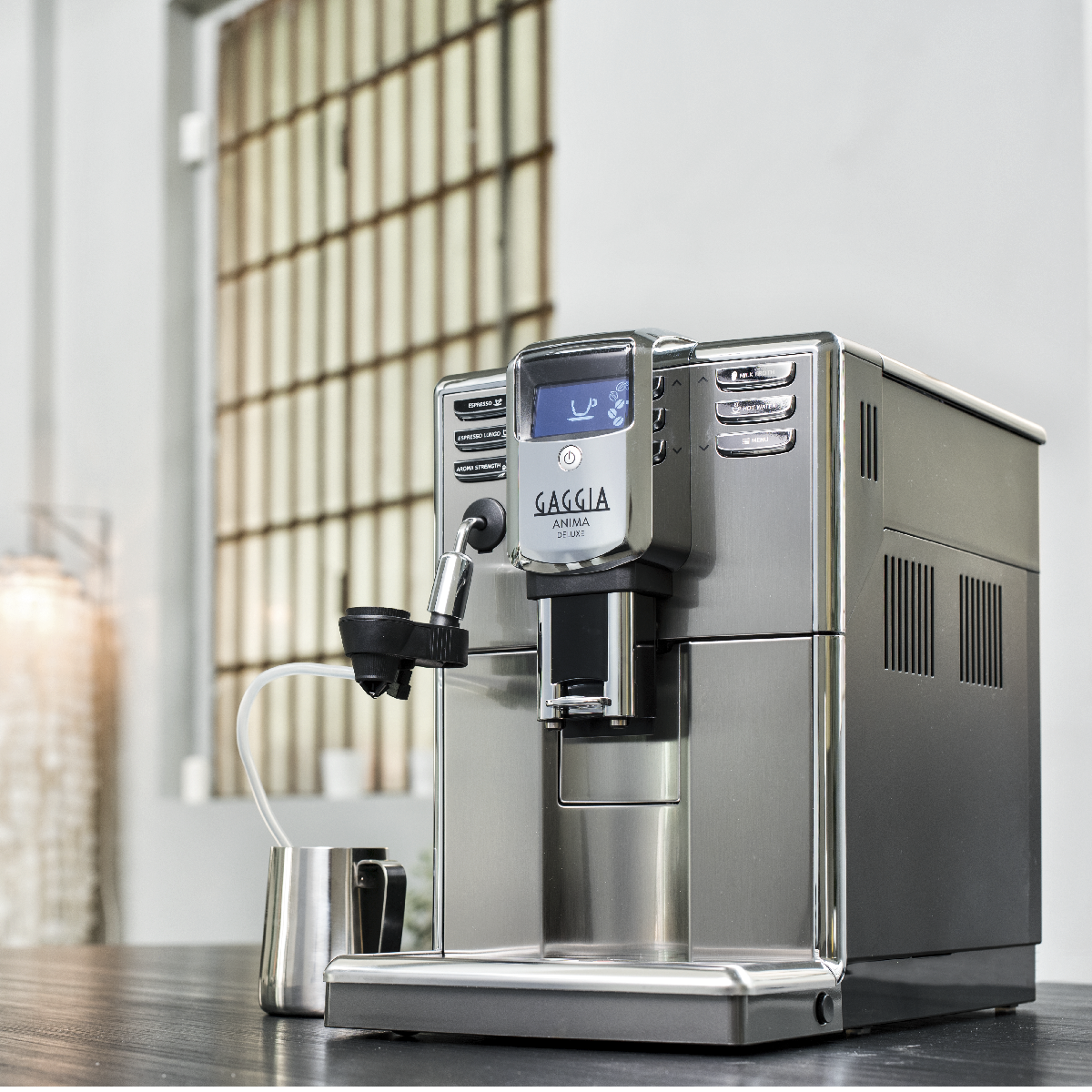 Ekspres do kawy Gaggia Anima De Luxe stoi na czarnej powierzchni. Mały dzbanek na mleko po lewej stronie. Okna w tle.