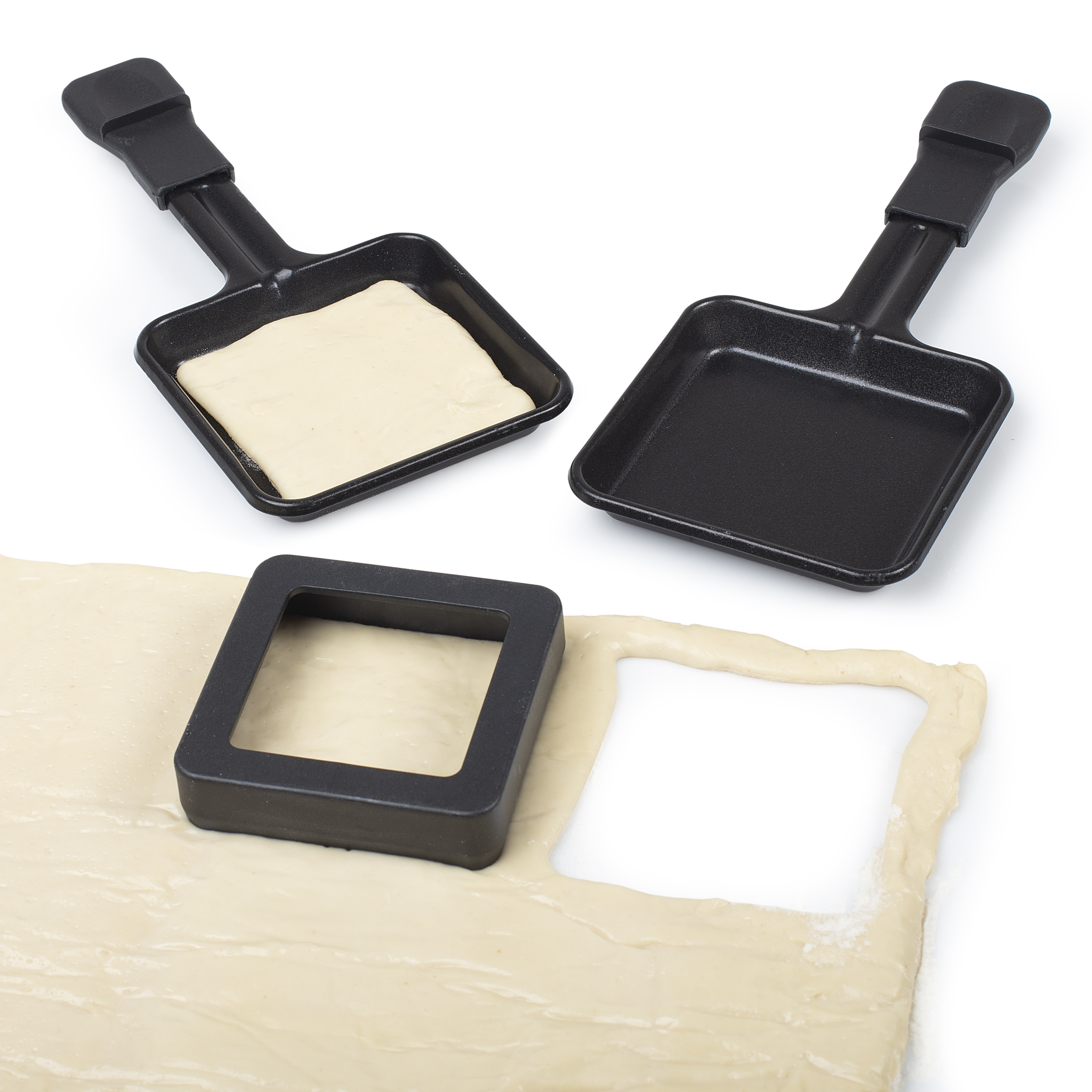 Dwie czarne patelnie z uchwytami, jedna wypełniona ciastem. Kwadratowy wykrawacz na cieście. Białe tło.