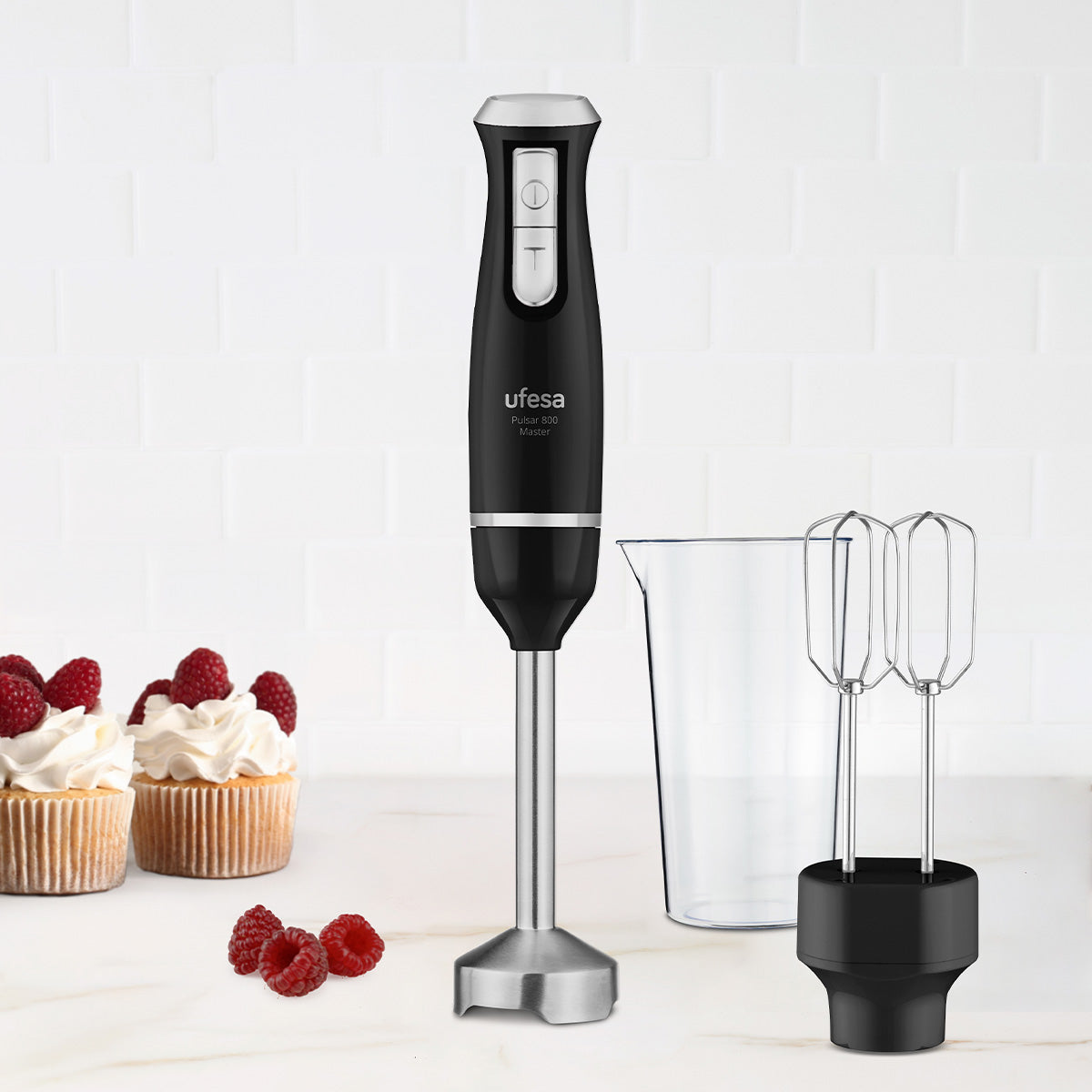 Un frullatore a immersione Ufesa nero, becher e accessori per mixer sono su un bancone. Due cupcake con lamponi sullo sfondo.