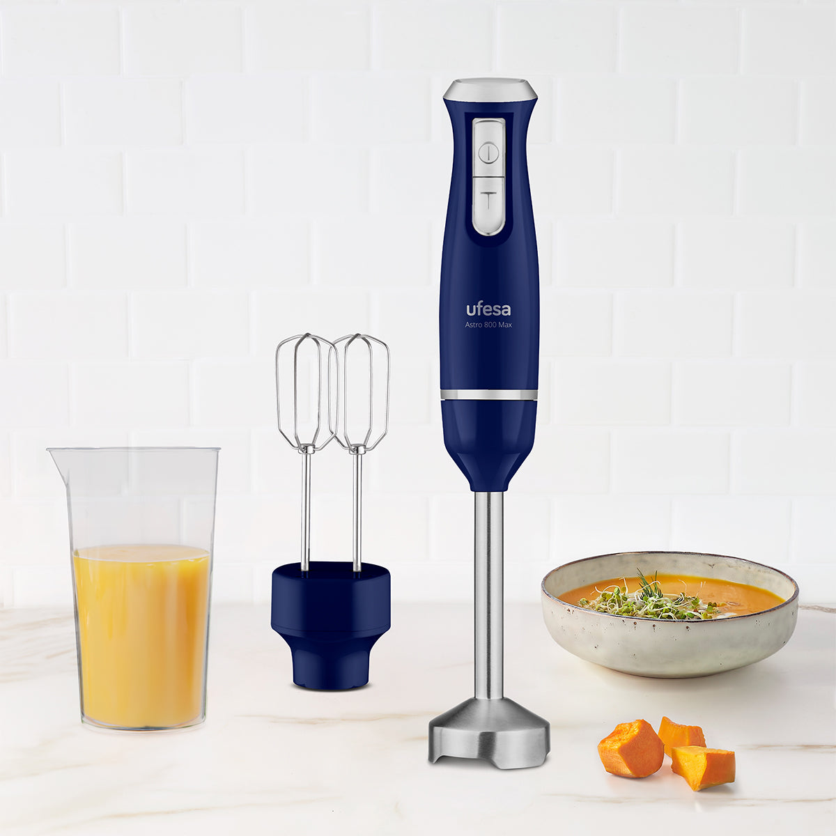 Un frullatore a immersione Ufesa blu con accessori, un bicchiere di liquido arancione, zuppa e zucca tritata su una superficie bianca.