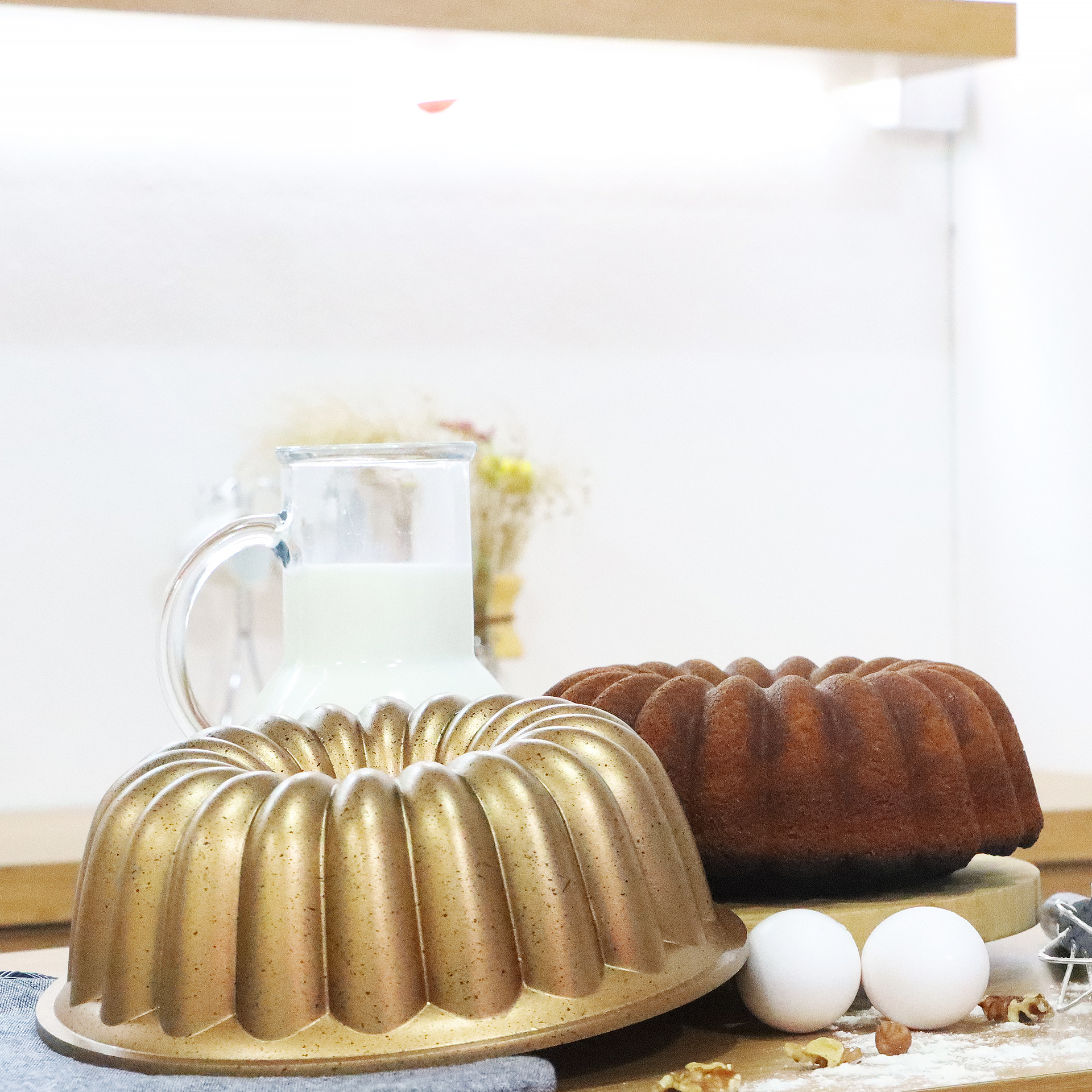 Un molde dorado para bizcocho junto a un pastel horneado, un vaso de leche, huevos y nueces sobre una mesa de madera.