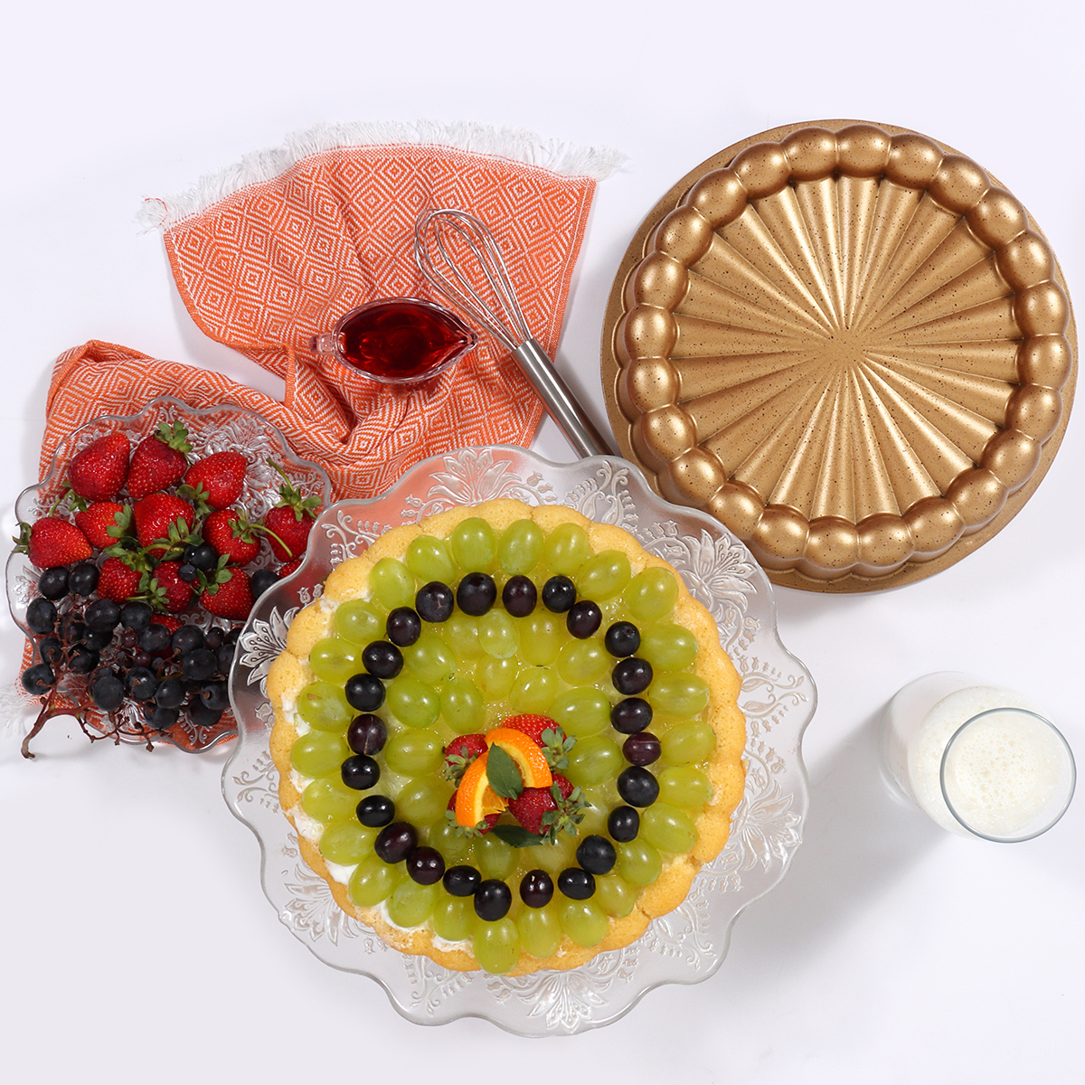 Tarta de frutas decorada con uvas verdes y negras, fresas y rodajas de naranja. Otros elementos incluyen una sartén dorada, vasos y un batidor.