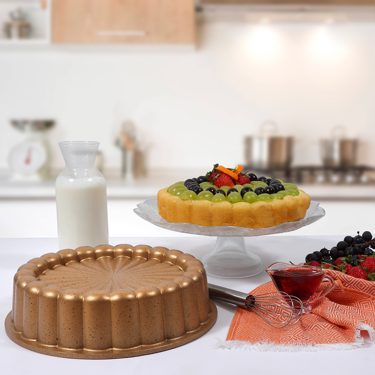 Una escena de cocina con un pastel en un soporte de vidrio, un molde para pastel, leche y fruta. Un cuenco con salsa, batidor y fruta.