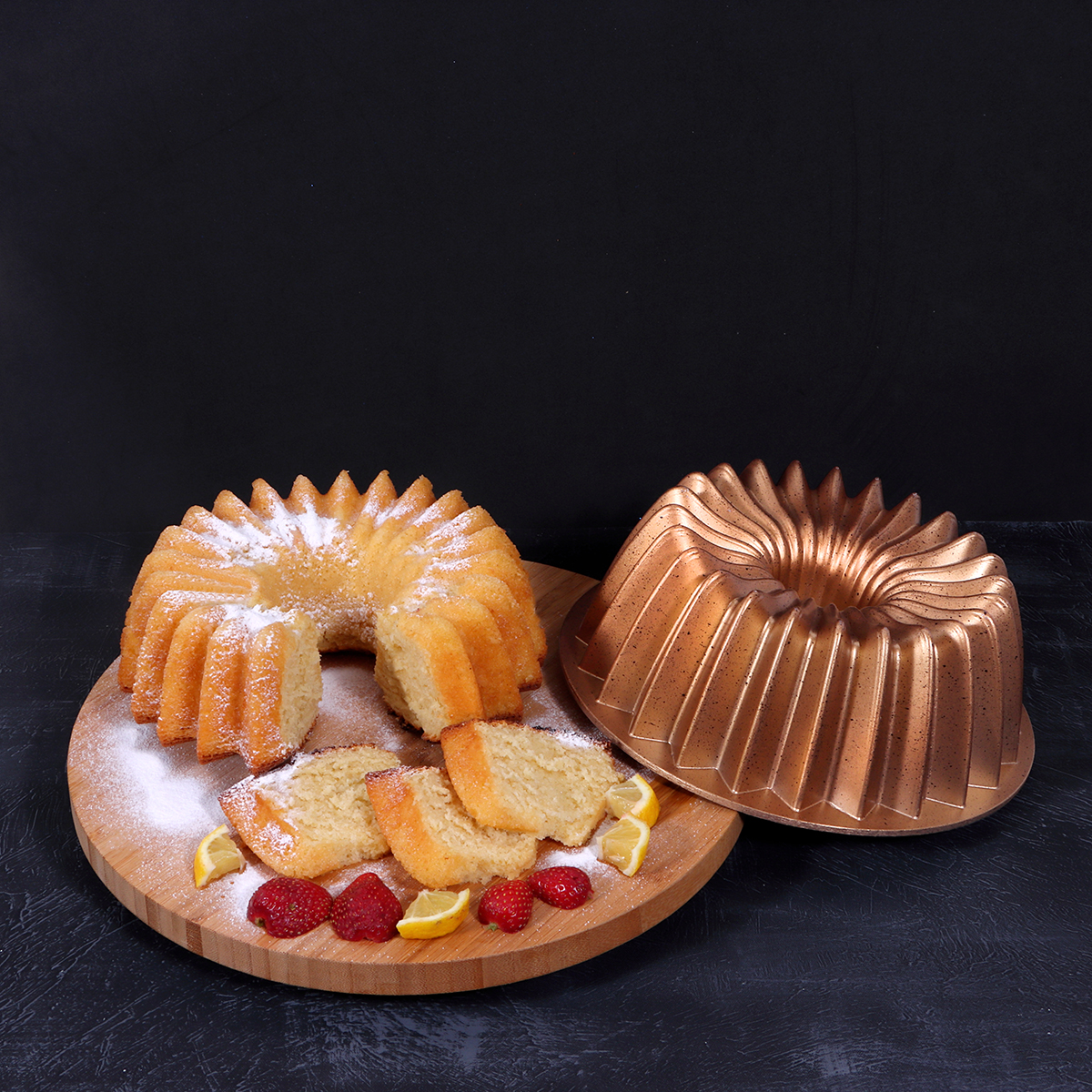 Un bizcocho con rebanadas, fresas y rodajas de limón sobre una tabla de madera. Una forma de bizcocho de cobre está al lado. Fondo negro.