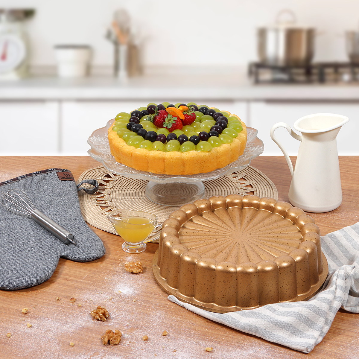 Un pastel de frutas con uvas y fresas está sobre un soporte de vidrio. Una tartera dorada y una jarra también están sobre la mesa.
