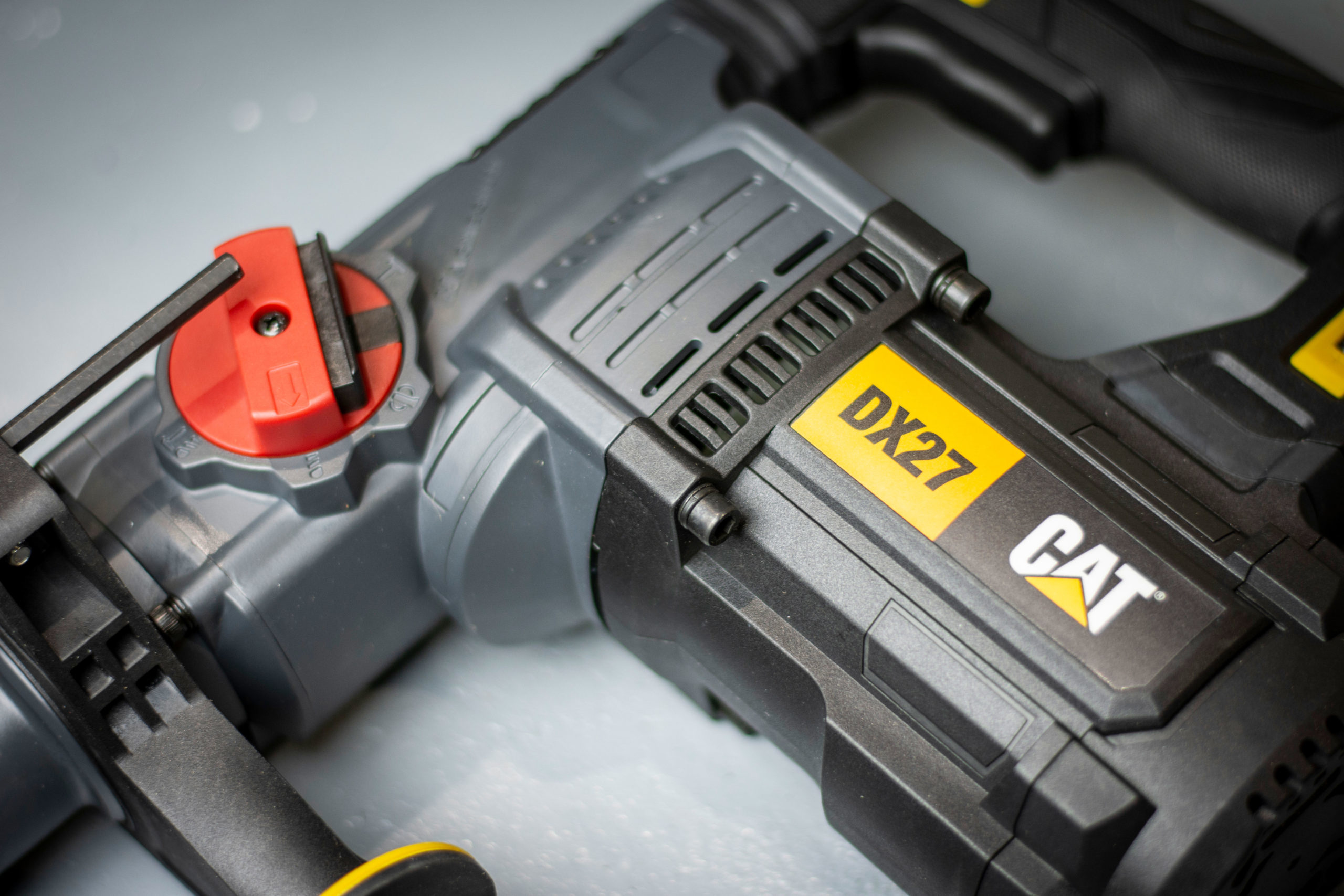 Nahaufnahme eines CAT DX27 Bohrhammers. Er hat einen roten Griff und gelbe Akzente. Die Bohrmaschine liegt auf grauem Untergrund.