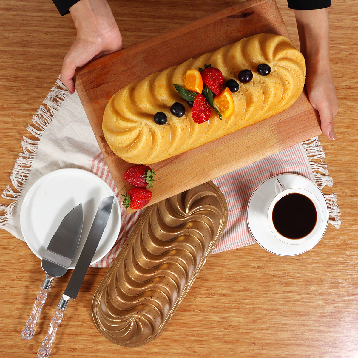 Un pastel en una tabla de madera decorado con frutas, sostenido por manos. Un plato, cuchillos, una taza de café y un molde para pastel están en la mesa.