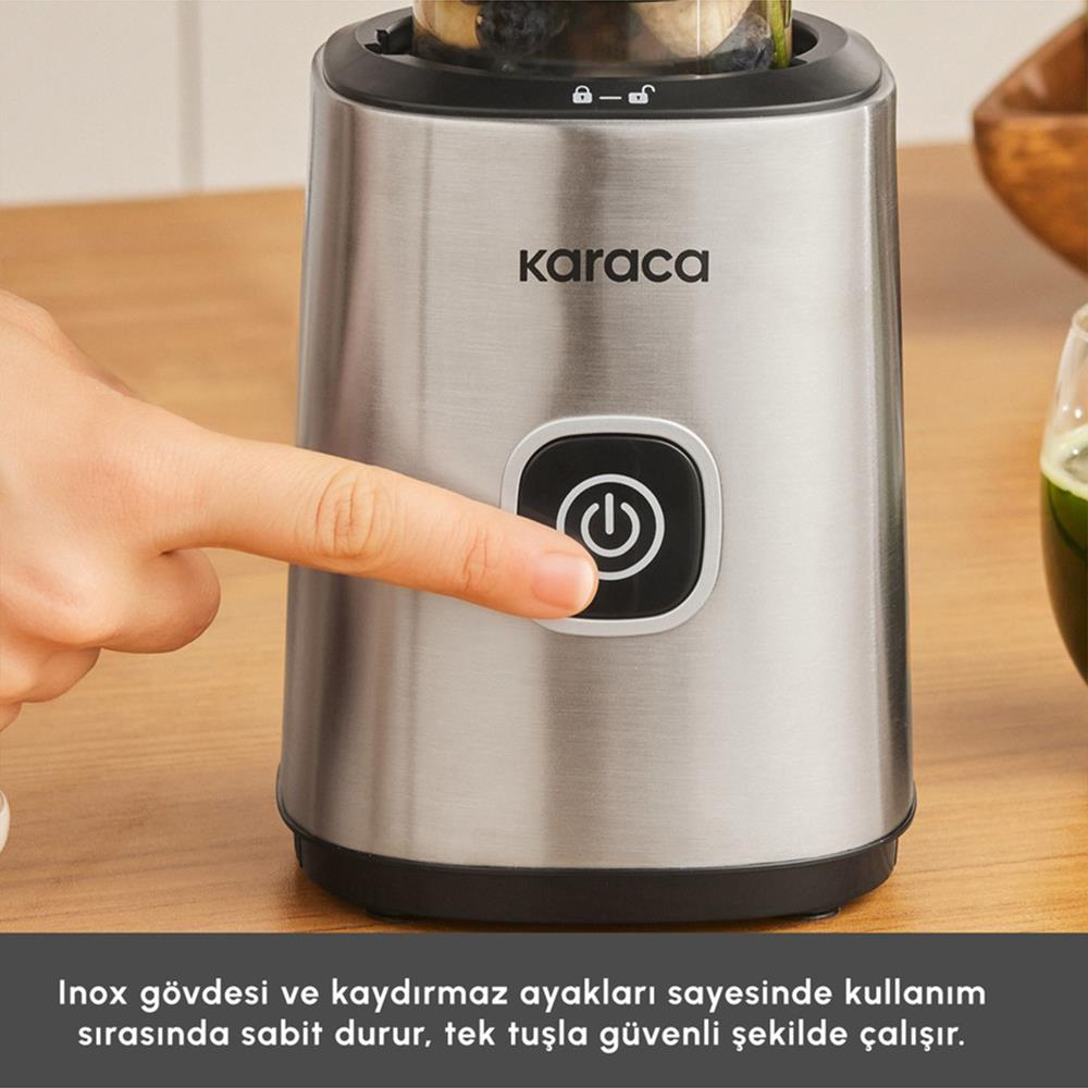 Bir el, gümüş renkli bir Karaca blenderin güç düğmesine basıyor. Arka planda ahşap bir yüzey ve bir bardak yeşil meyve suyu görülüyor.
