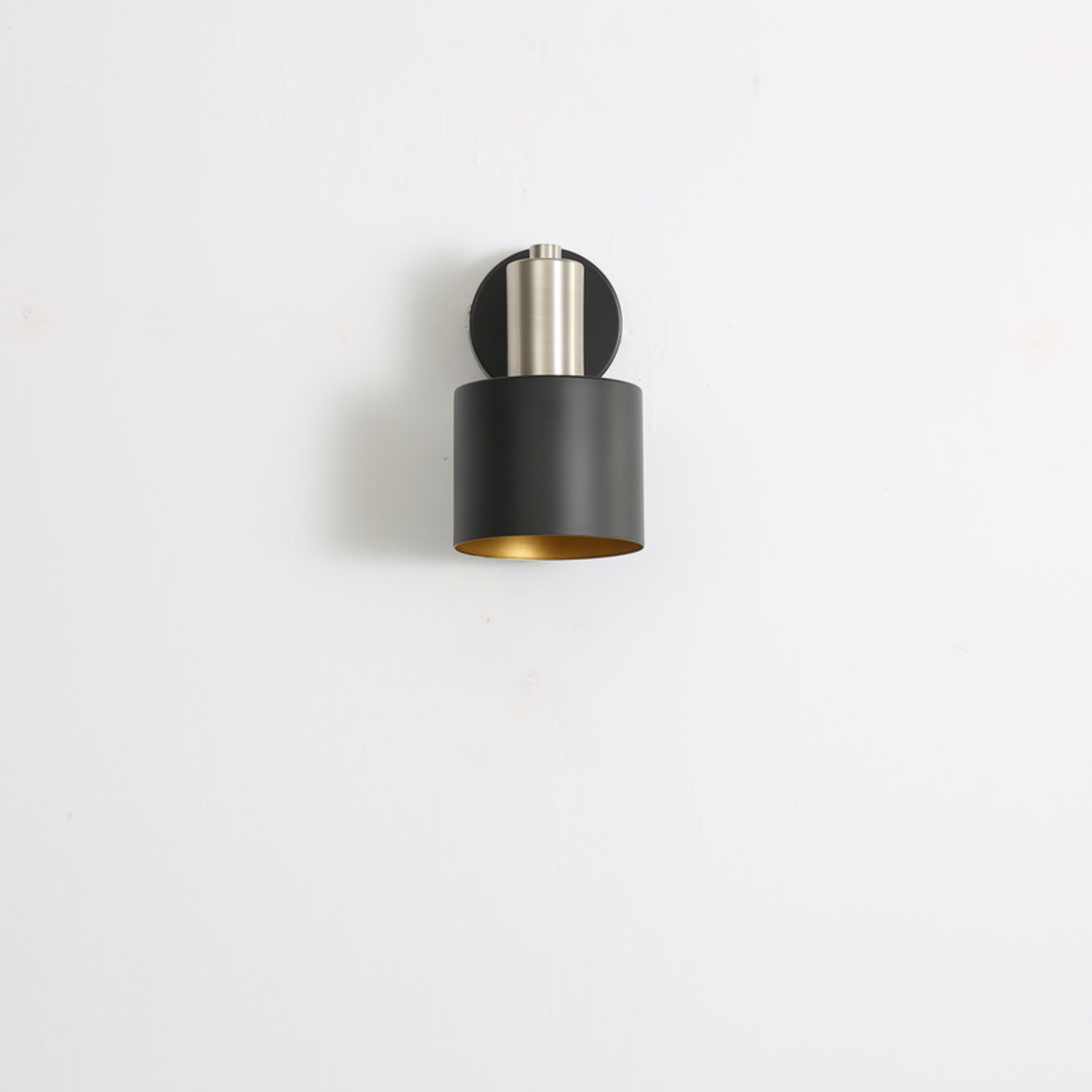 Una moderna lampada da parete nera con interno dorato, montata su un muro bianco. La lampada ha un paralume cilindrico.