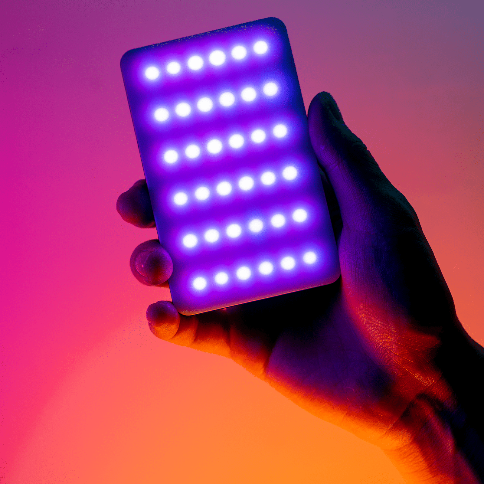 Hand holding a rectangular device with bright white lights on a gradient pink to orange background.