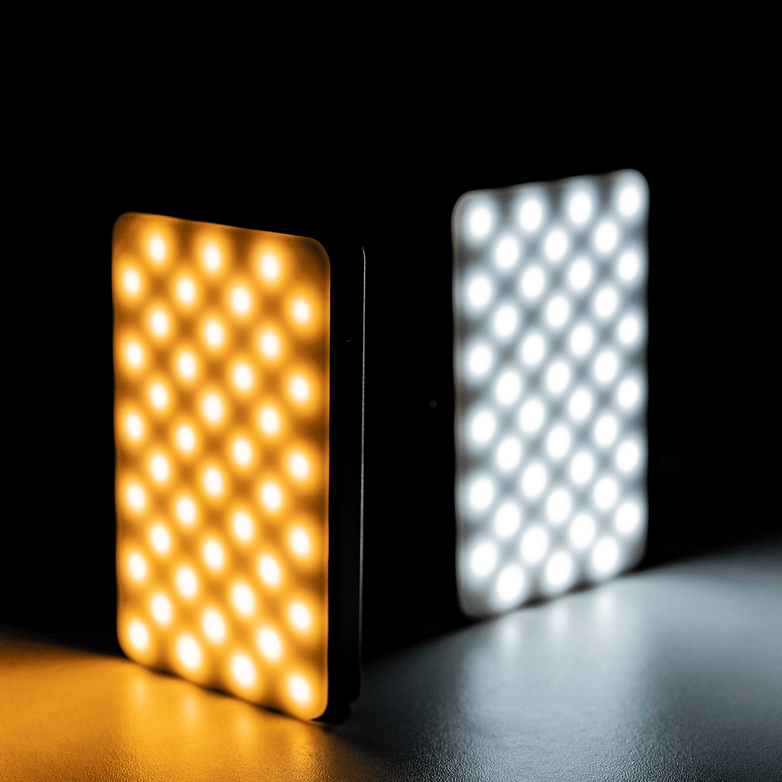 Two LED panels, one warm, one cool, side by side, illuminated. Dark background and surface create high contrast.