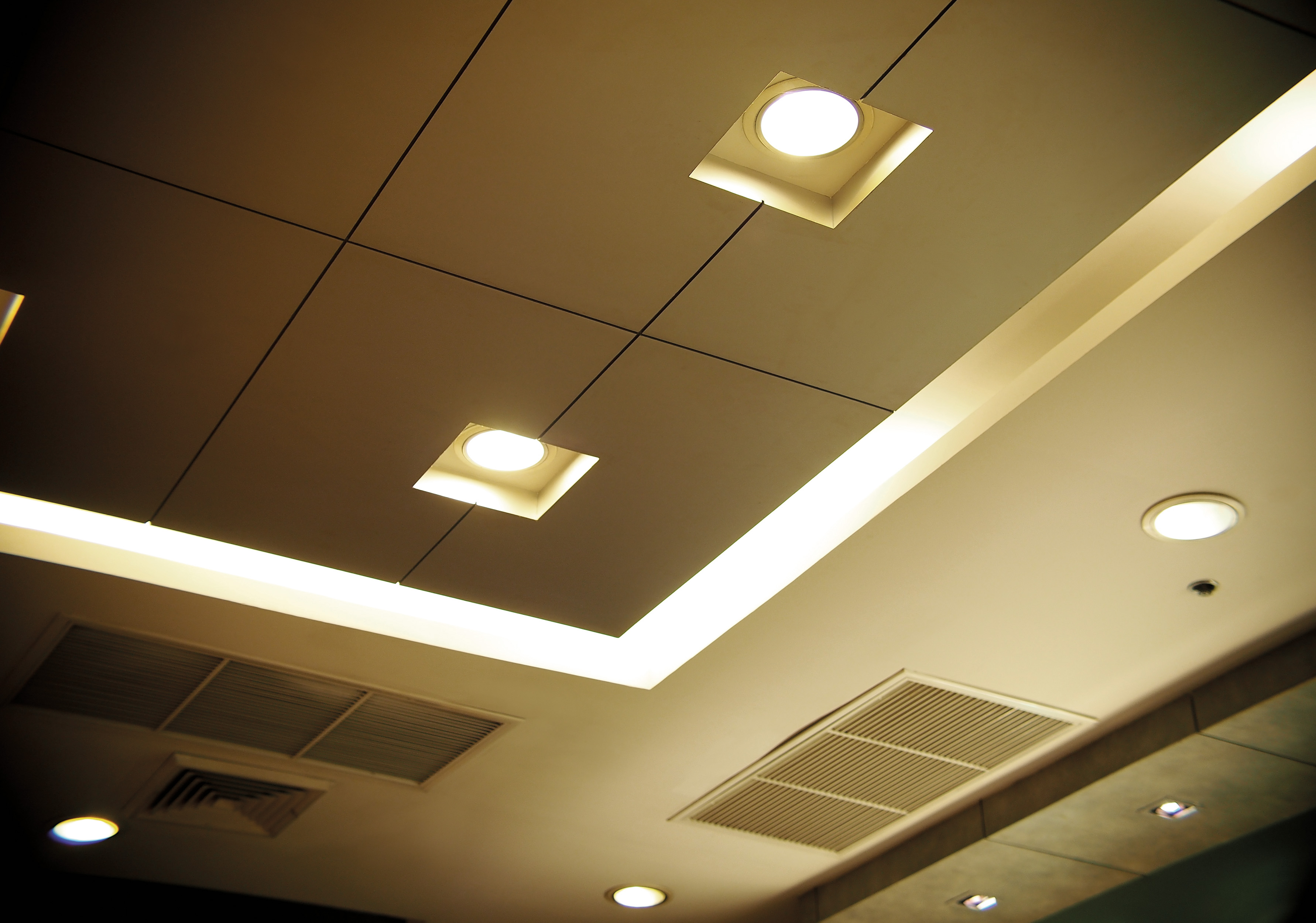 Eine Decke mit Lichtern und Lüftungsschlitzen ist zu sehen. Die Decke hat quadratische und runde Lichter. Die Farbe ist gelblich.