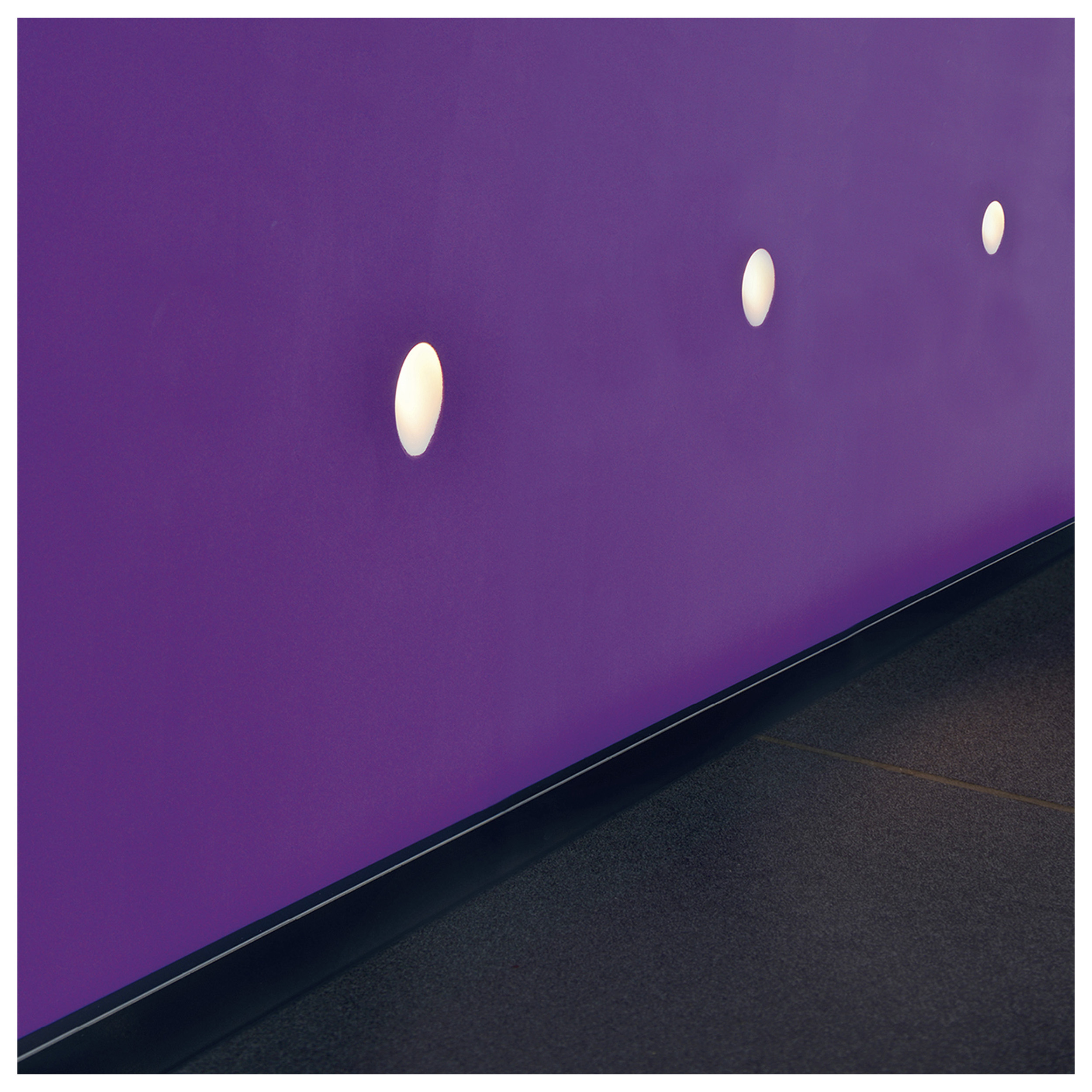 Eine lila Wand mit ovalen Lichtern. Darunter ein dunkler Boden. Wand und Boden treffen sich an einer schwarzen Kante.