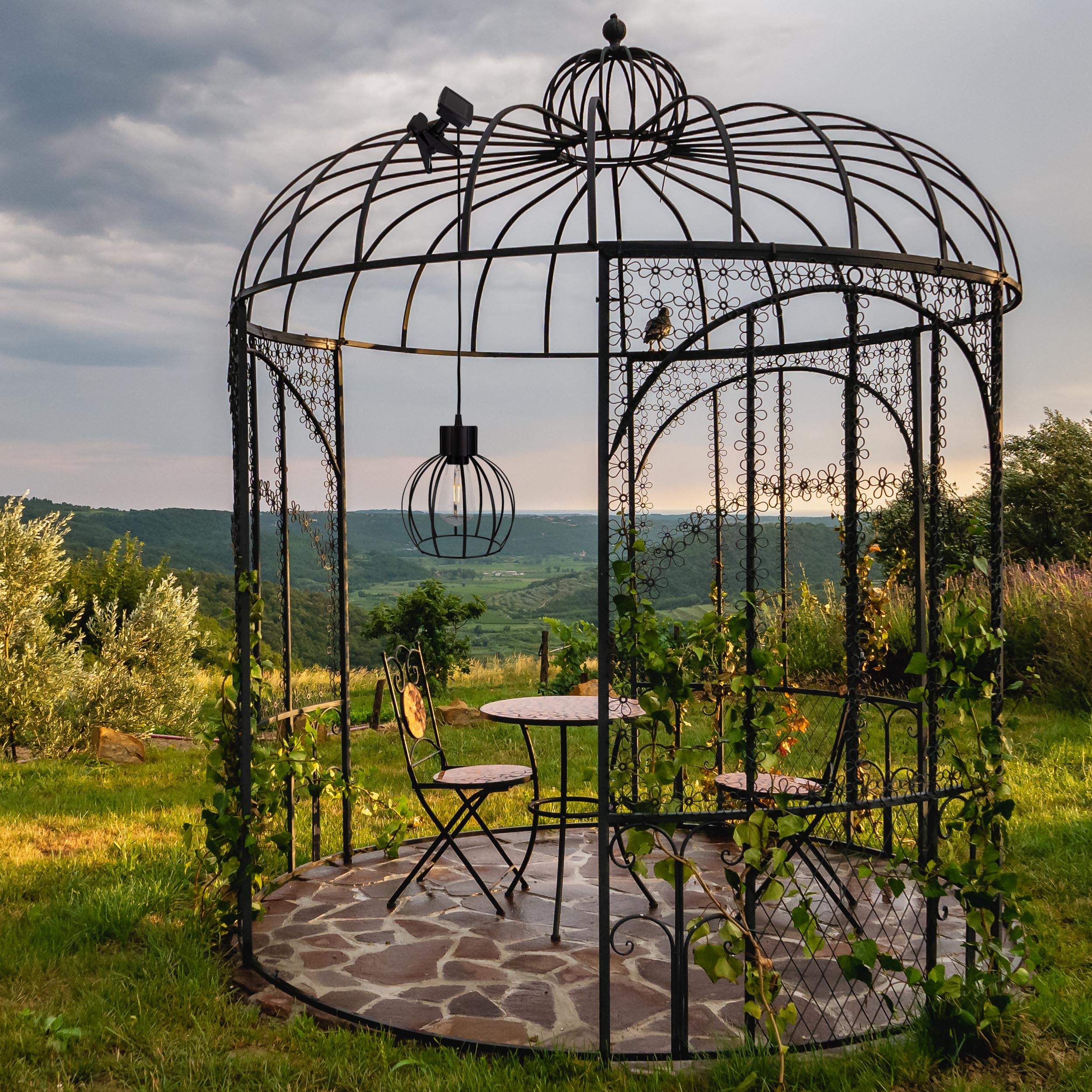 Ein Pavillon mit Tisch und Stühlen steht auf einer Wiese mit Blick auf ein Tal. Ein Vogel sitzt auf dem Pavillon.