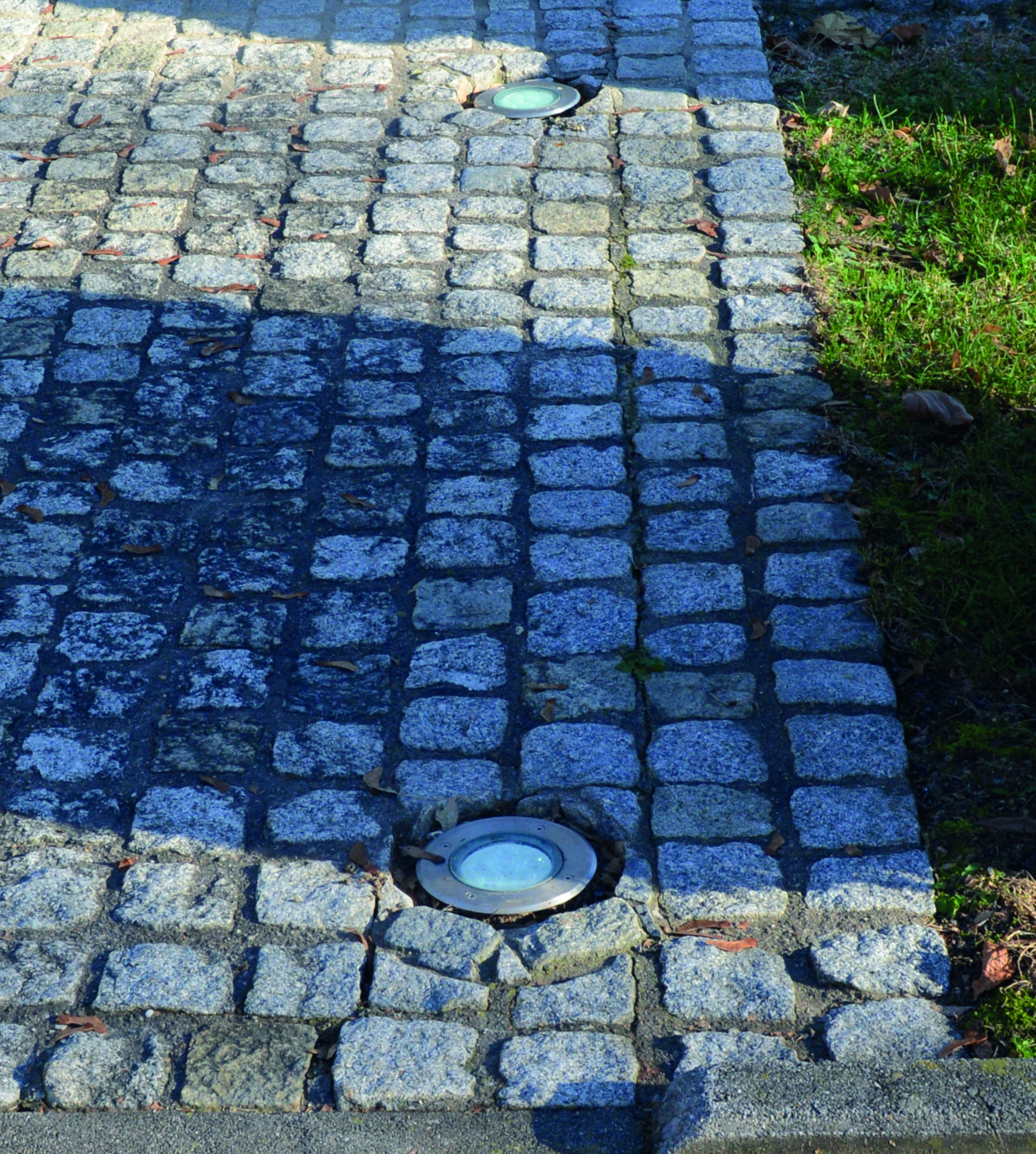 Steinweg mit drei eingebetteten Lichtern. Rechts Gras und Blätter. Der Weg besteht aus grauen Steinen.