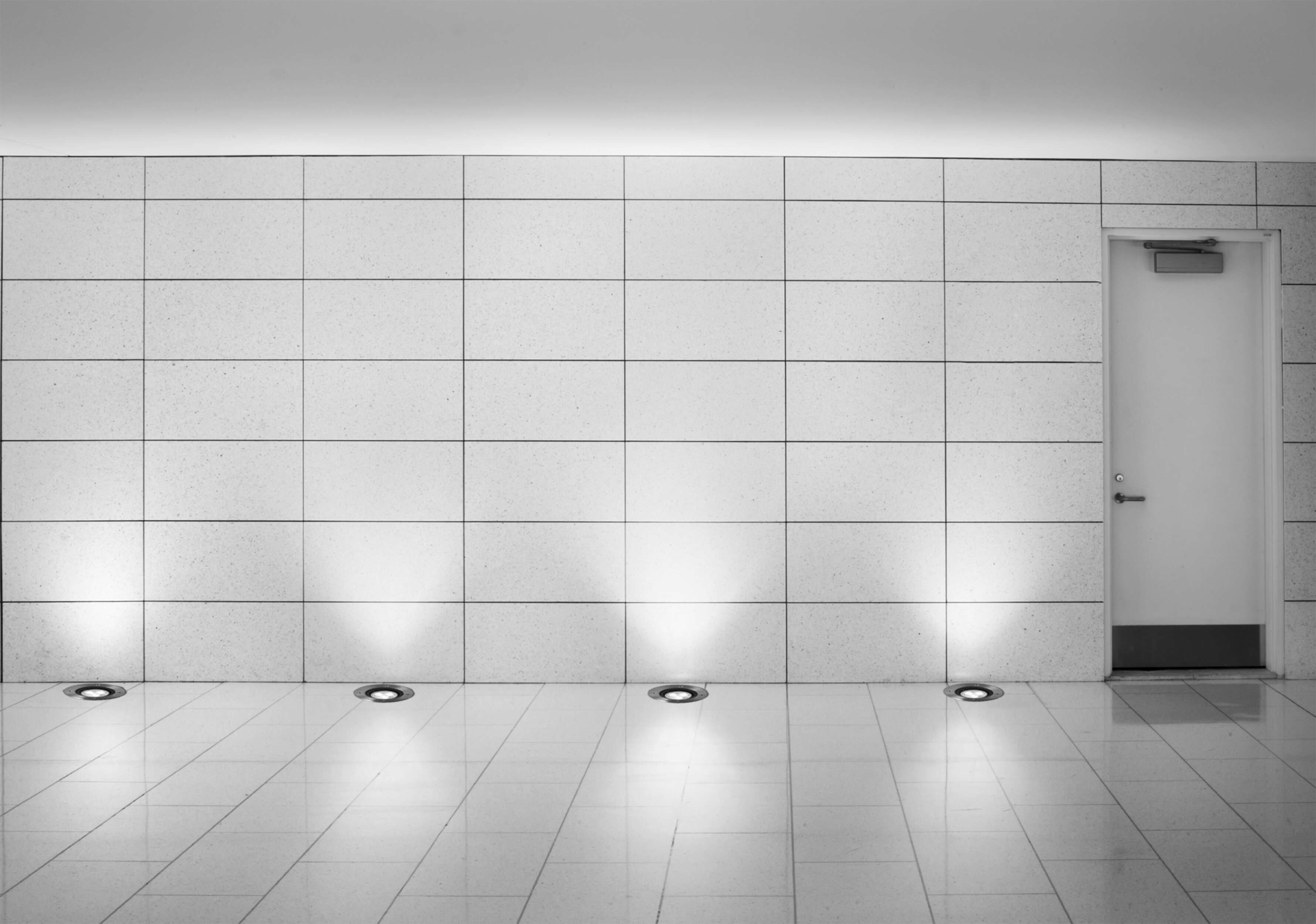 Monochromes Interieur. Geflieste Wand mit Tür rechts. Bodenleuchten beleuchten den Boden. Weiße Tür.