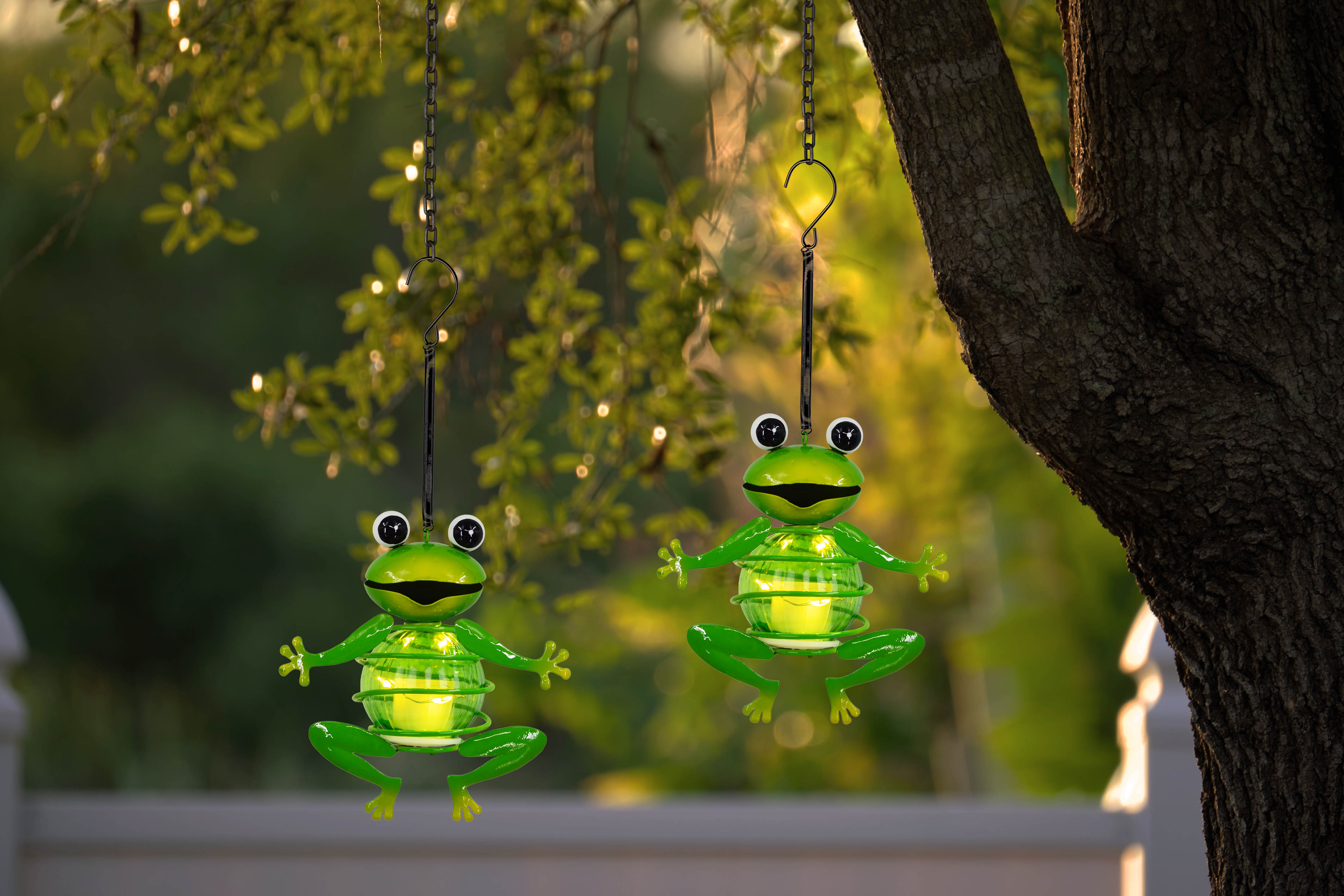 Zwei grüne Froschdekorationen hängen an Ketten. Die Frösche sind in der Nähe eines Baumes. Der Hintergrund ist verschwommen.
