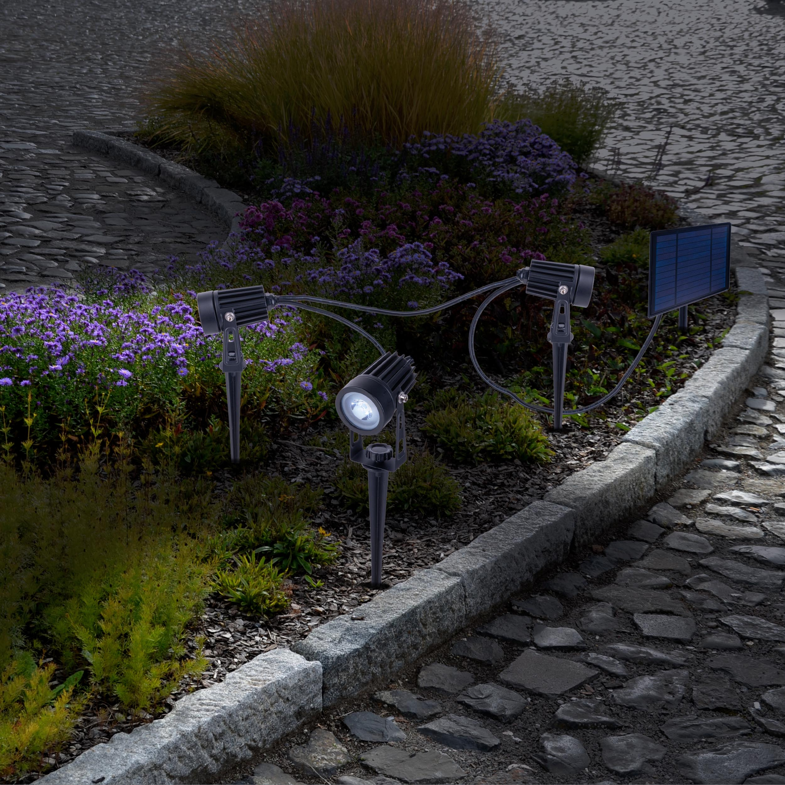 Garten-Strahler beleuchten lila Blumen und Grün. Kopfsteinpflaster und Solarpanel sind ebenfalls sichtbar.