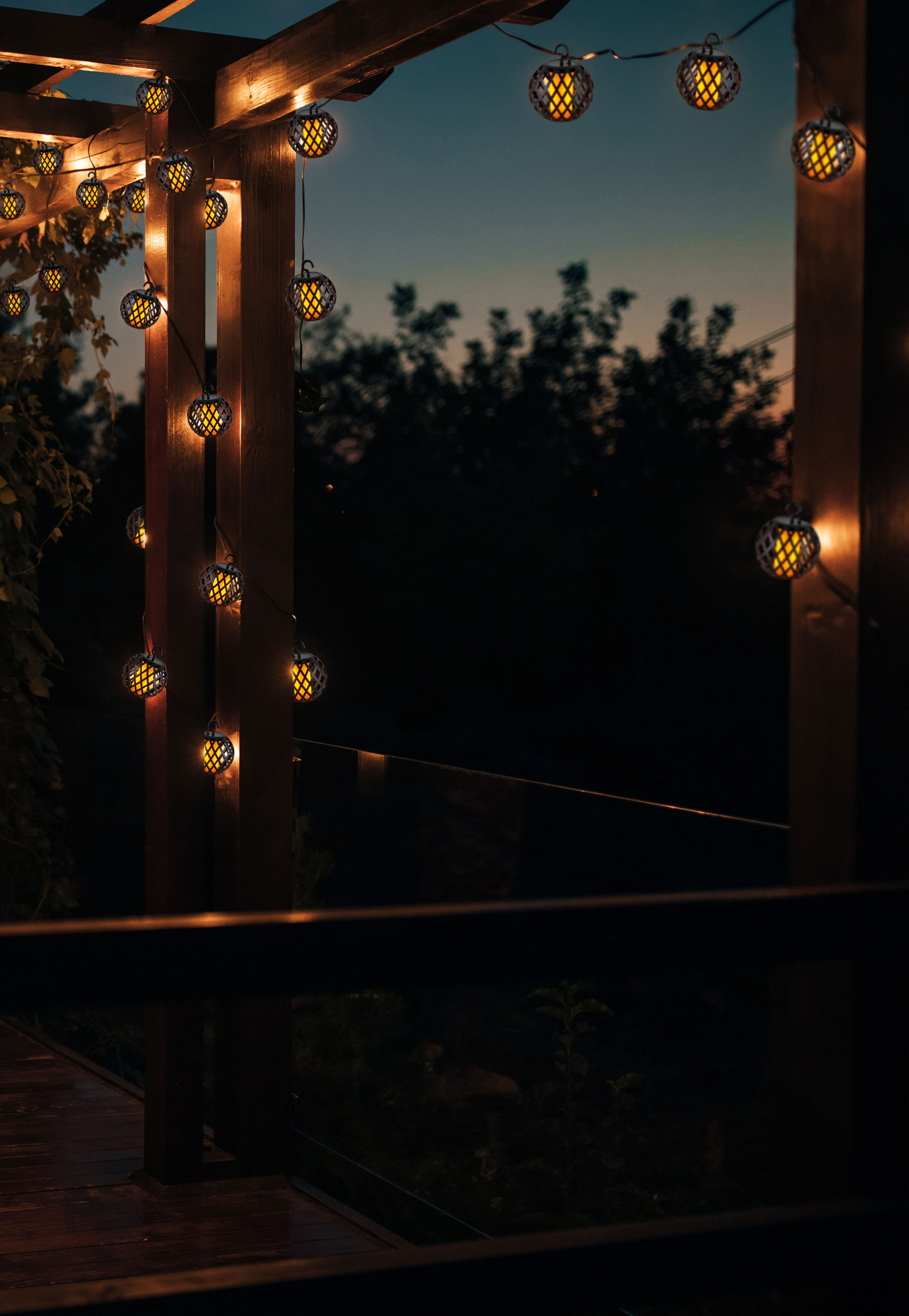 Lichterketten mit gelbem Schein hängen von einer Holzpergola in der Abenddämmerung. Dunkle Bäume bilden den Hintergrund.