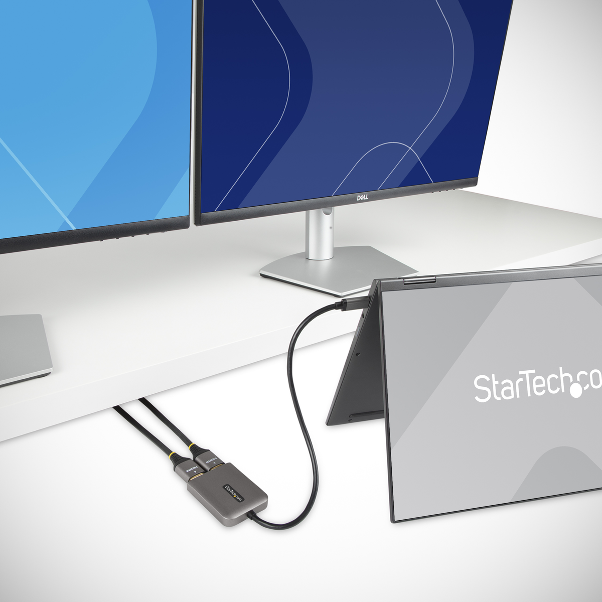 Due monitor e un laptop collegati a un dispositivo grigio. Gli schermi mostrano disegni blu e blu scuro. Logo StarTech.com visibile.