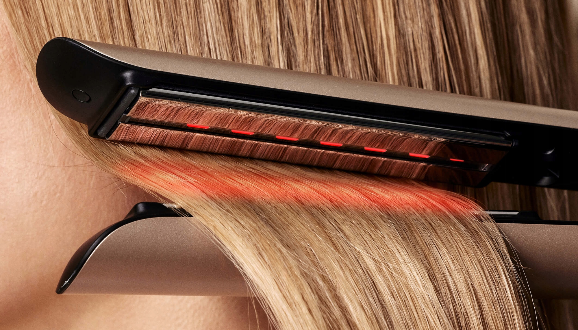 Ein Glätteisen glättet blondes Haar. Das Eisen ist gold-schwarz mit roten Lichtern. Haare sind im Vorder- und Hintergrund.