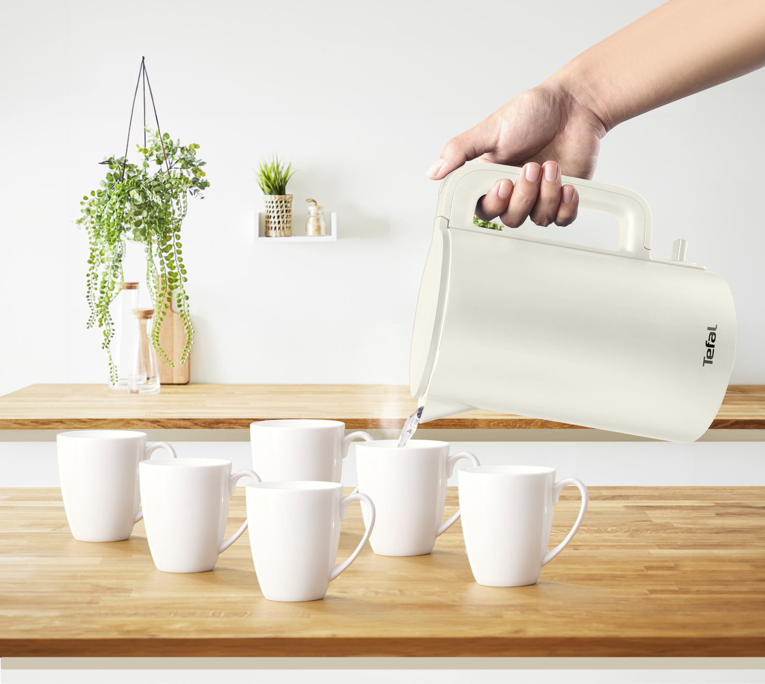 Une main verse de l'eau d'une bouilloire crème dans des tasses blanches sur une surface en bois. Plantes en arrière-plan.