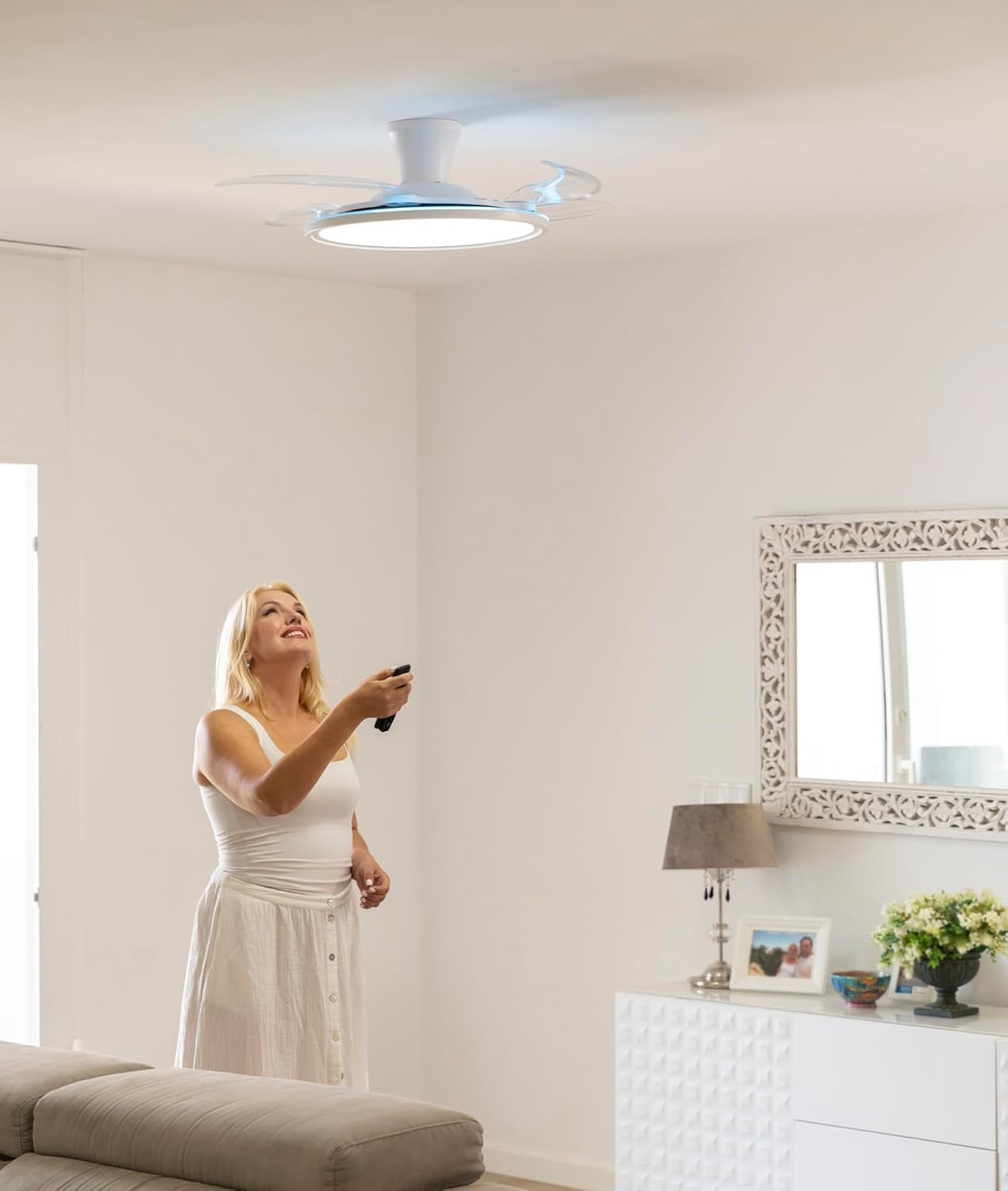 Mujer con vestido blanco mira ventilador de techo. Sostiene mando. Habitación blanca con sofá, espejo, lámpara, flores y mueble blanco.