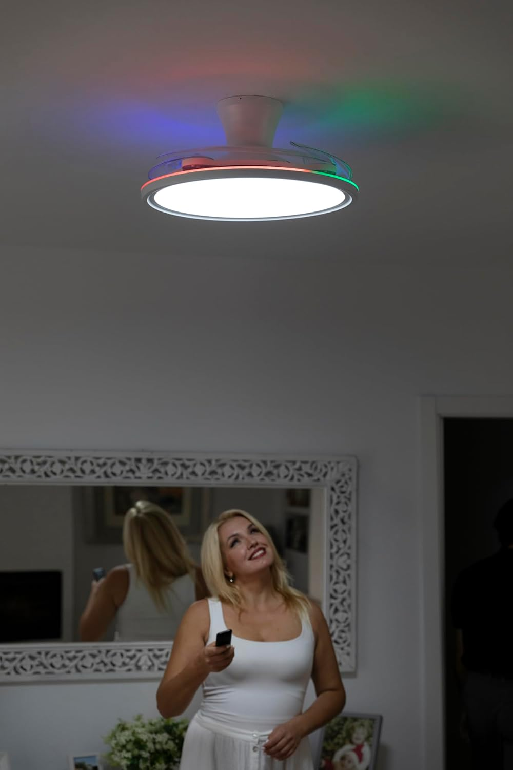 Una mujer mira hacia arriba a un ventilador de techo con luces de colores. Sostiene un mando a distancia, cerca de un espejo que la refleja.