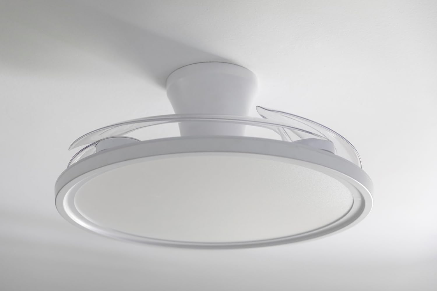 Un ventilador de techo blanco con aspas transparentes está montado en un techo blanco. El ventilador tiene una lámpara circular.