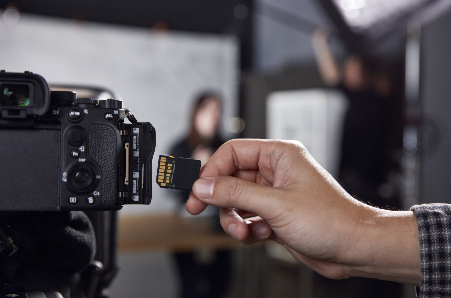 Ręka wkłada kartę pamięci do aparatu. Aparat jest czarny. Rozmyte postacie w tle.
