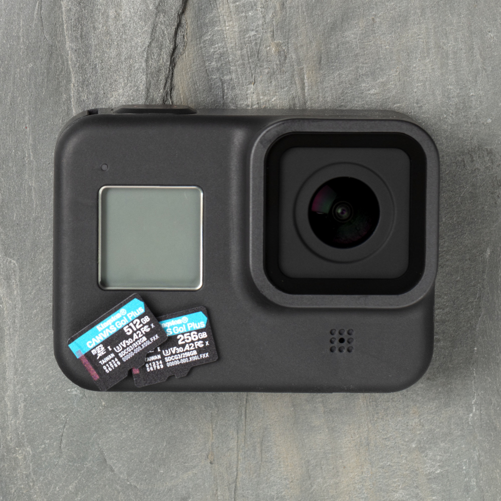 Zwarte actiecamera met twee SD-kaarten op een grijze stenen ondergrond. De camera heeft een scherm en een lens. De kaarten hebben tekst.