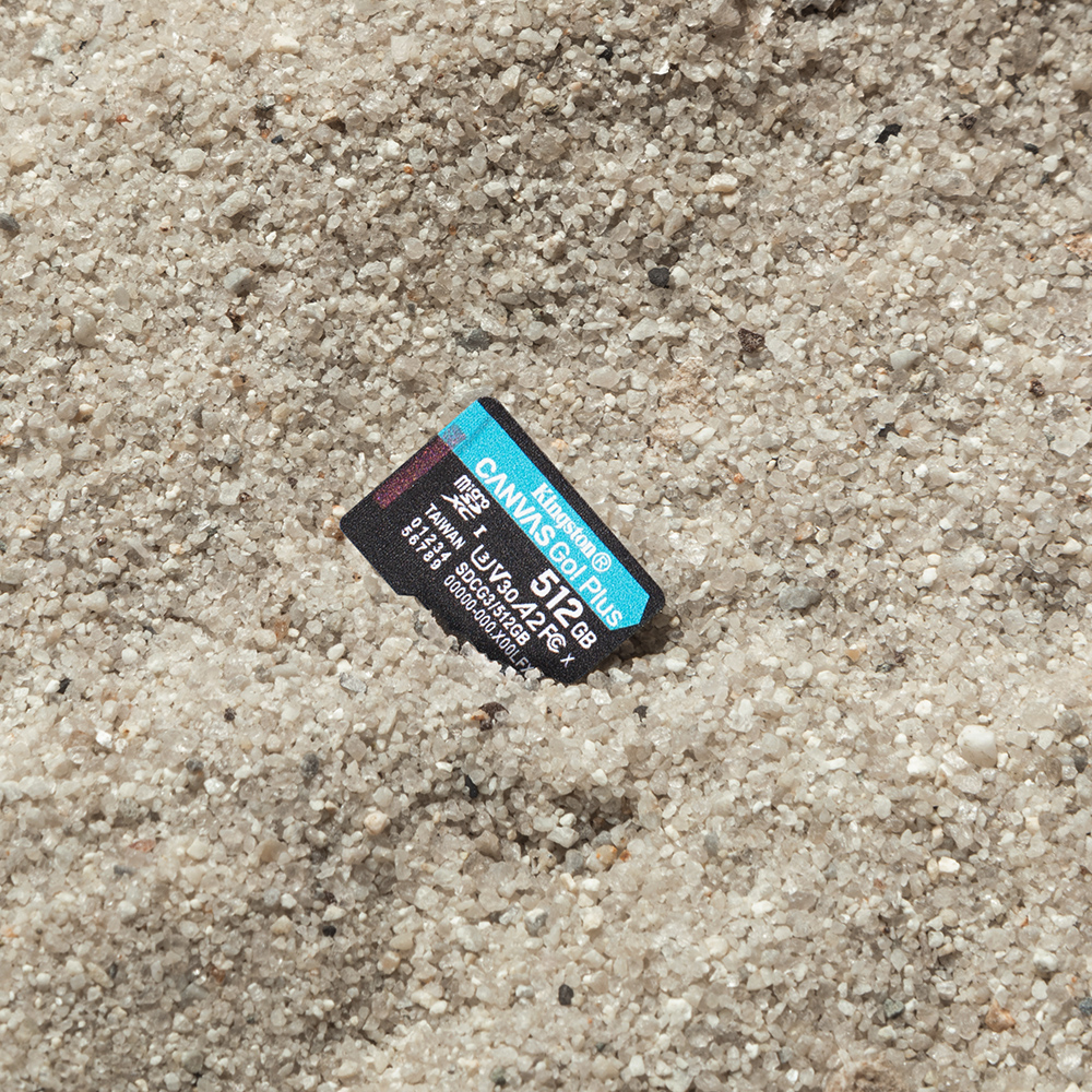Een Kingston Canvas Go! Plus 512 GB micro SD-kaart ligt op zand. De kaart is zwart, turquoise en wit.