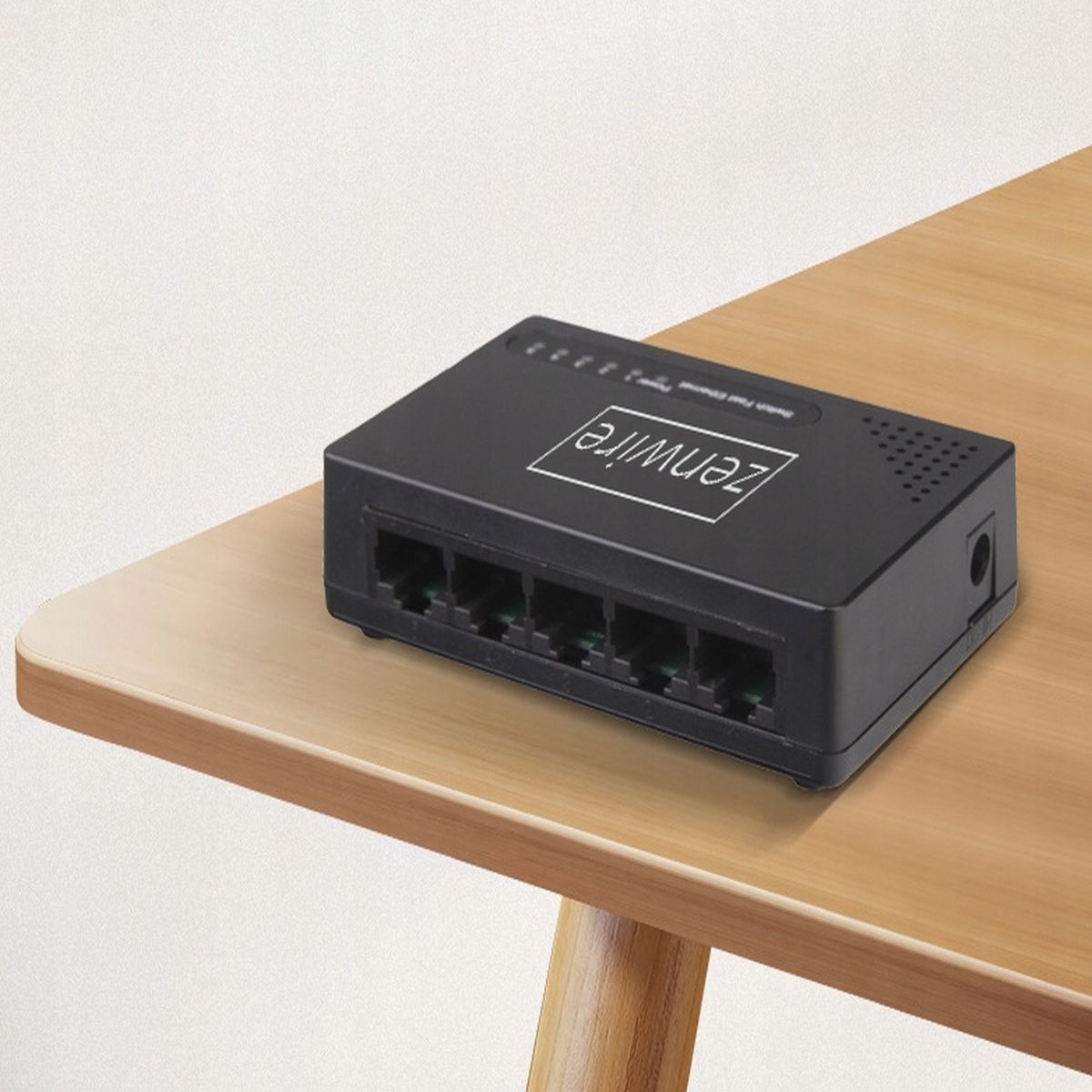 Czarny przełącznik sieciowy z napisem 'Zenwire' leży na jasnym drewnianym stole. Stół ma zaokrągloną krawędź i widoczną nogę.