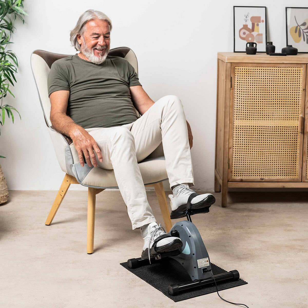 Un hombre mayor con una camisa verde y pantalones blancos usa una mini bicicleta de ejercicios. Está en una habitación con un armario de madera.