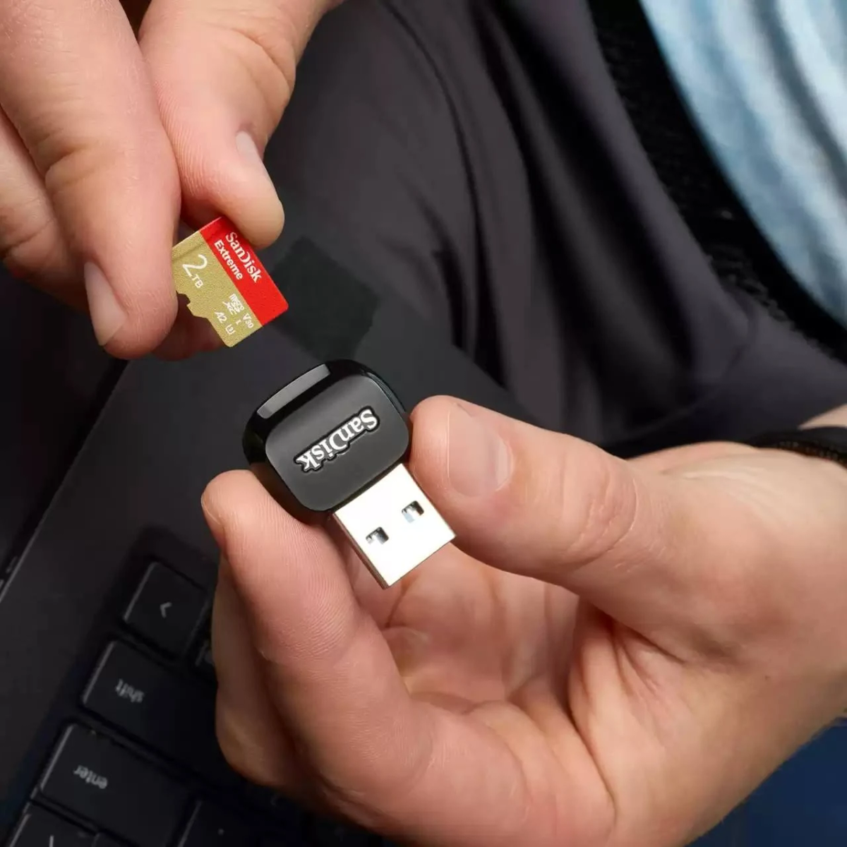 Manos sosteniendo una tarjeta micro SD Sandisk roja y dorada sobre un adaptador USB negro, junto a una laptop.