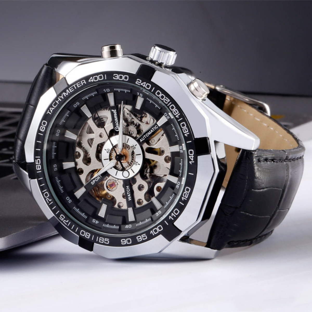 Nahaufnahme einer silber-schwarzen Armbanduhr mit sichtbarem Mechanismus auf weißem Untergrund, mit schwarzem Lederarmband.