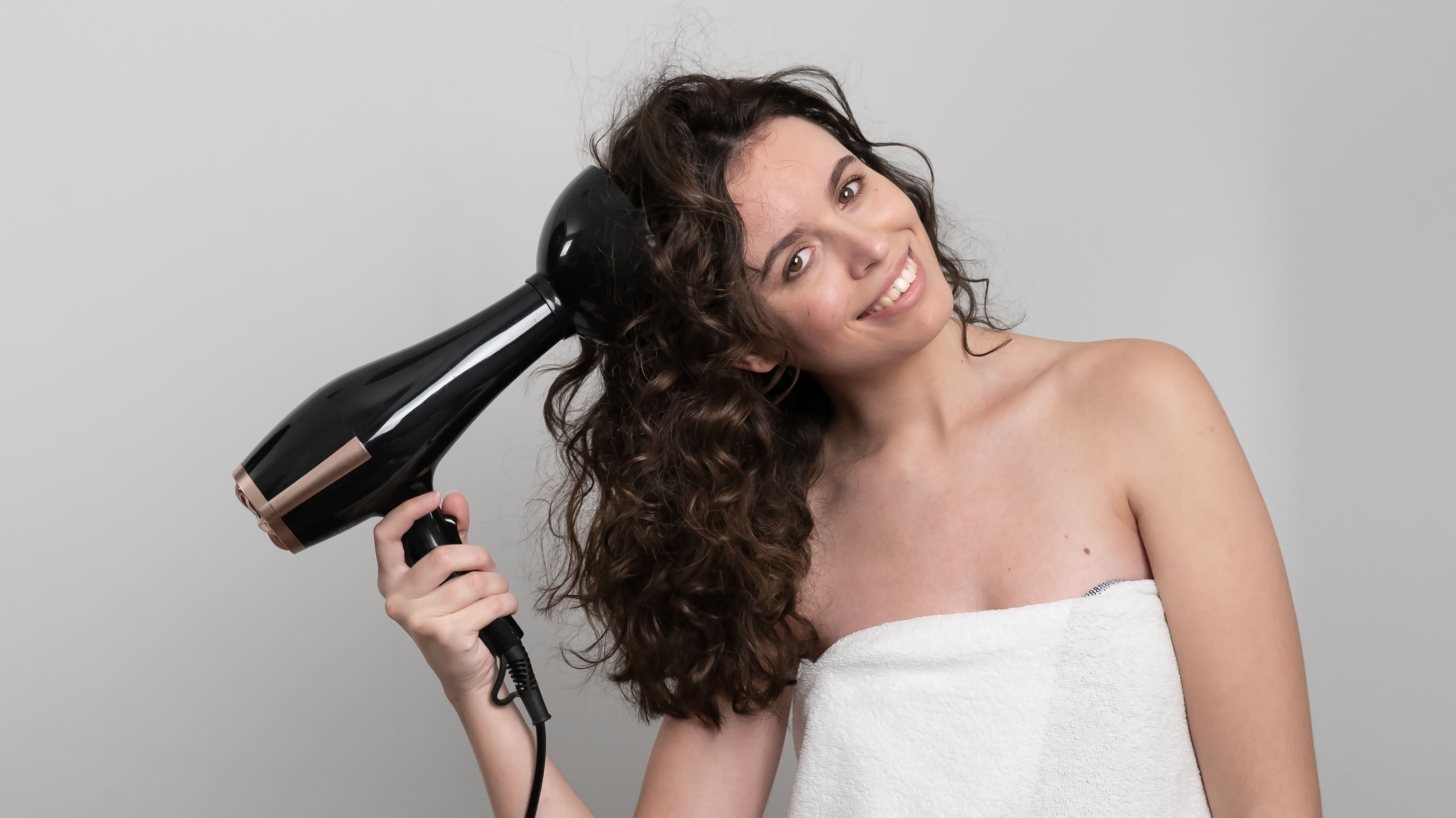 Una donna asciuga i suoi capelli ricci con un asciugacapelli nero. Sorride, avvolta in un asciugamano bianco, su sfondo grigio.