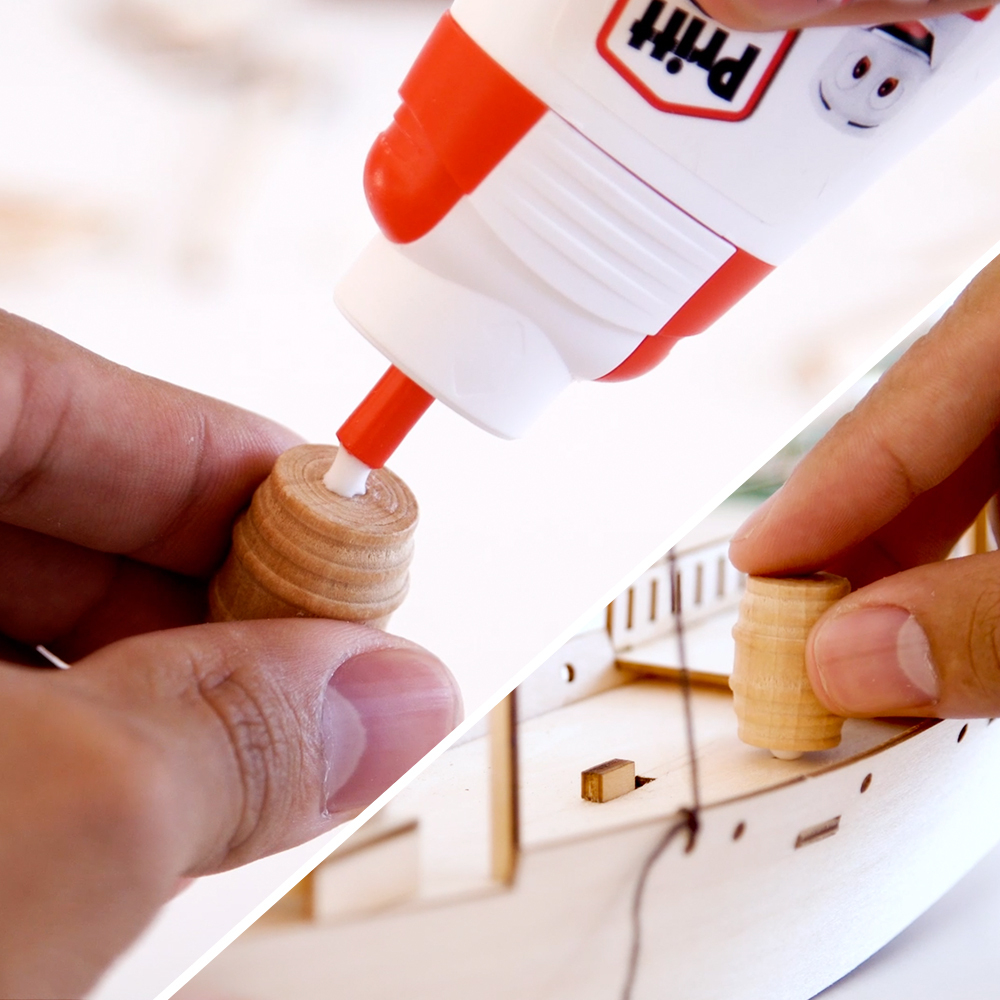 Eine Person trägt Kleber aus einer Pritt-Flasche auf ein kleines Holzfass auf. Ein weiteres Fass wird auf ein Holzschiffsmodell gelegt.
