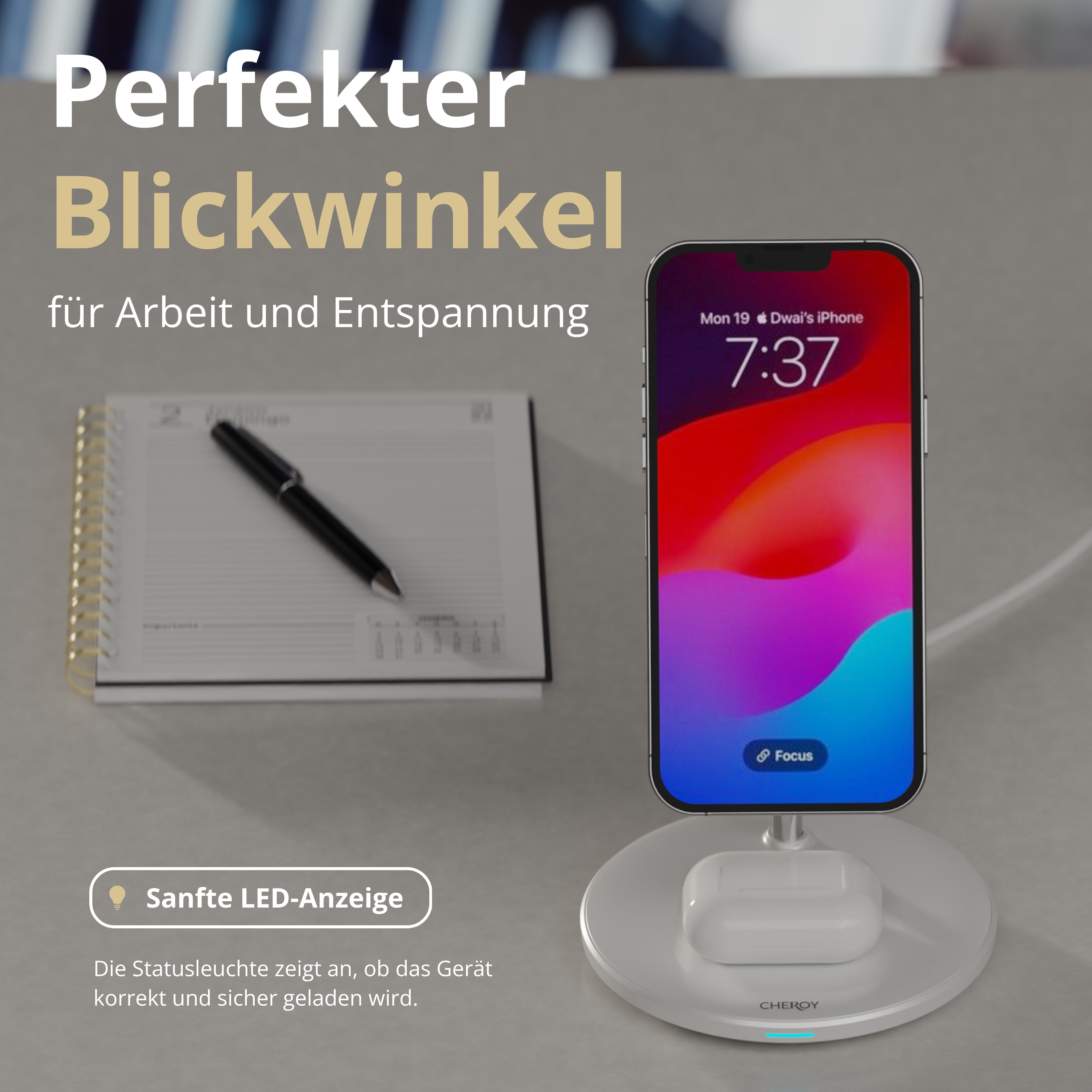 Ein iPhone auf einem Ladegerät, ein Notizbuch mit einem Stift und Text auf Deutsch auf einer hellgrauen Oberfläche.
