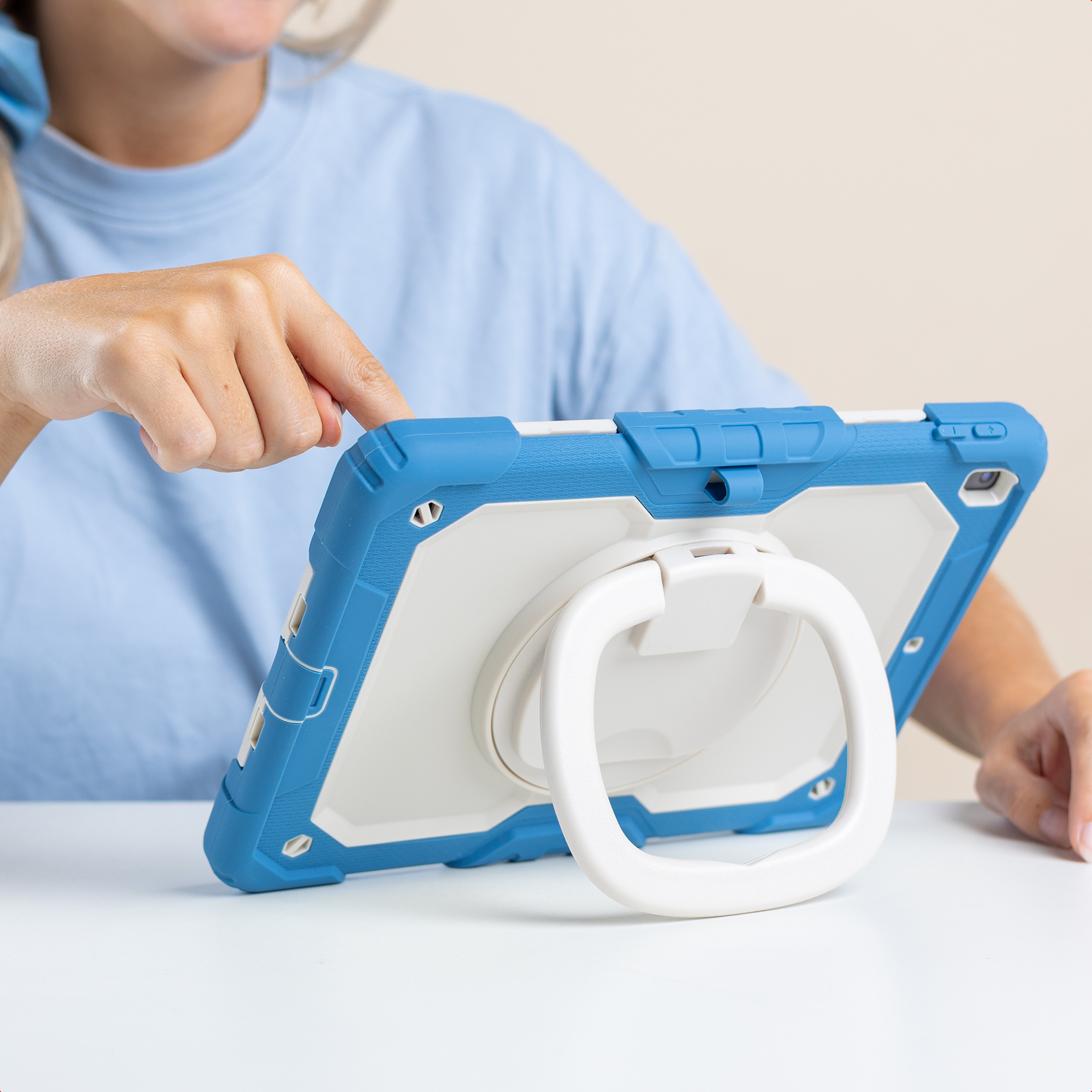 Une personne utilise une tablette bleue et blanche avec un support. La tablette est sur une table blanche. Le fond est crème.