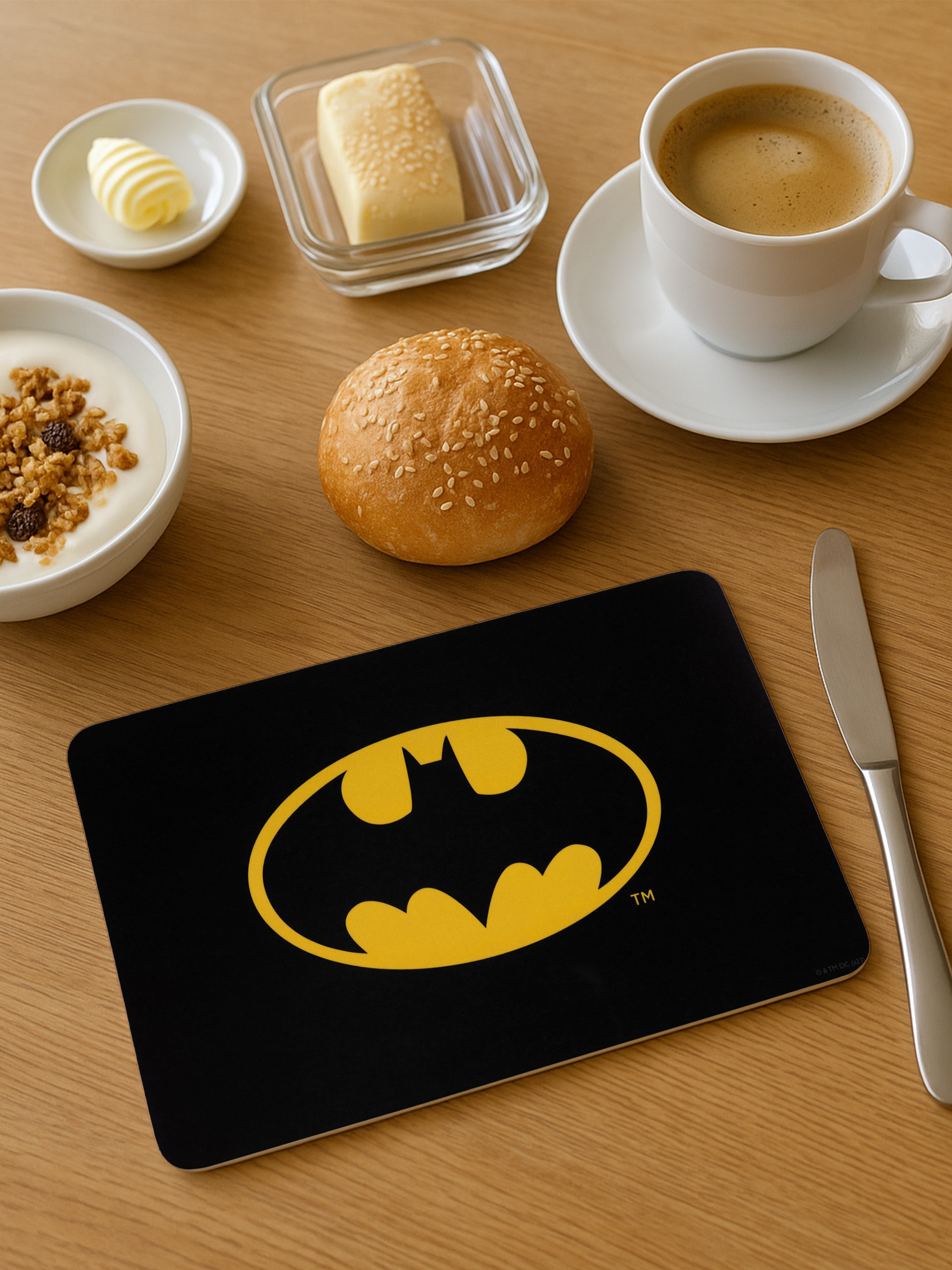 Frühstücksszene: Kaffee, Butter, Brötchen, Joghurt und eine Batman-Logo-Tischmatte auf Holz.