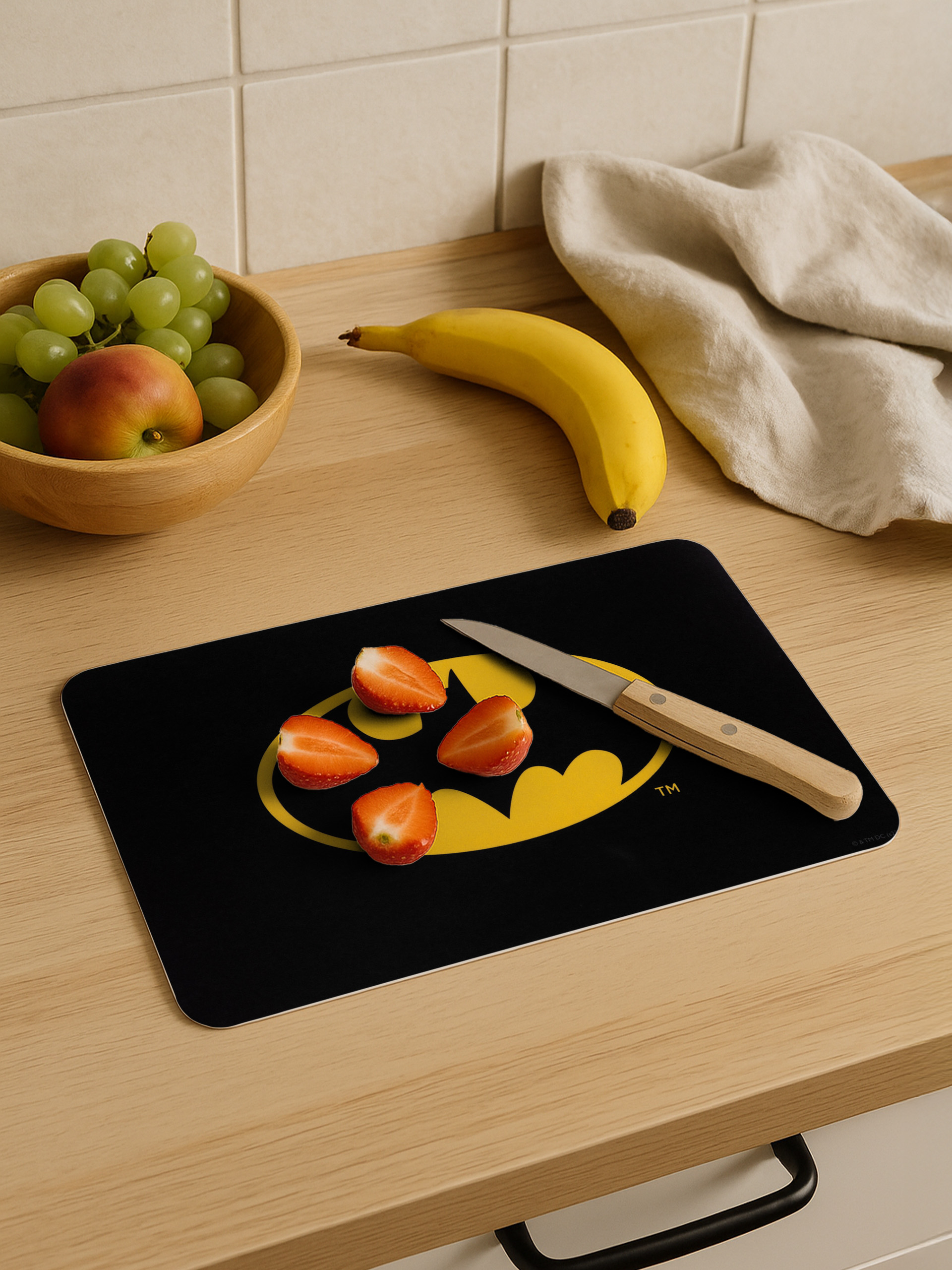 Küchenszene mit Erdbeeren auf einem Batman-Logo-Schneidebrett, Banane, Trauben und einem Messer.