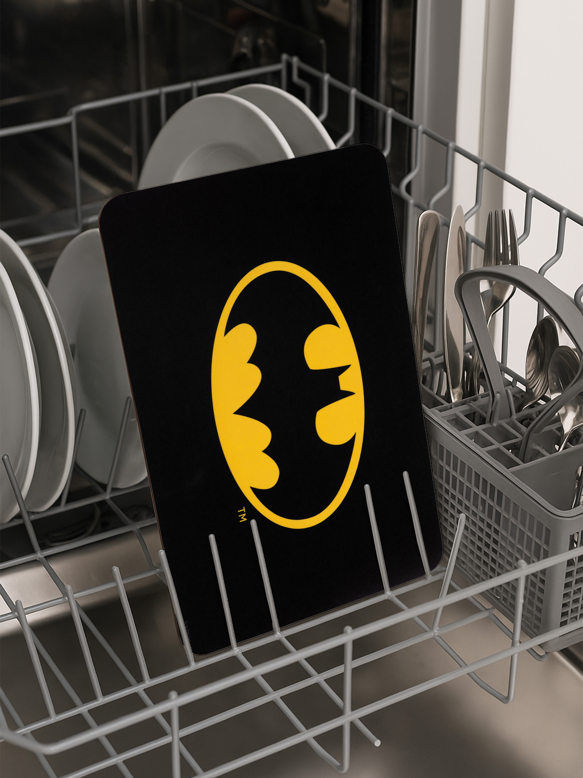 Eine Batman-Logo-Platte in einer Spülmaschine. Das Logo ist gelb auf schwarz. Andere Teller und Besteck sind sichtbar.