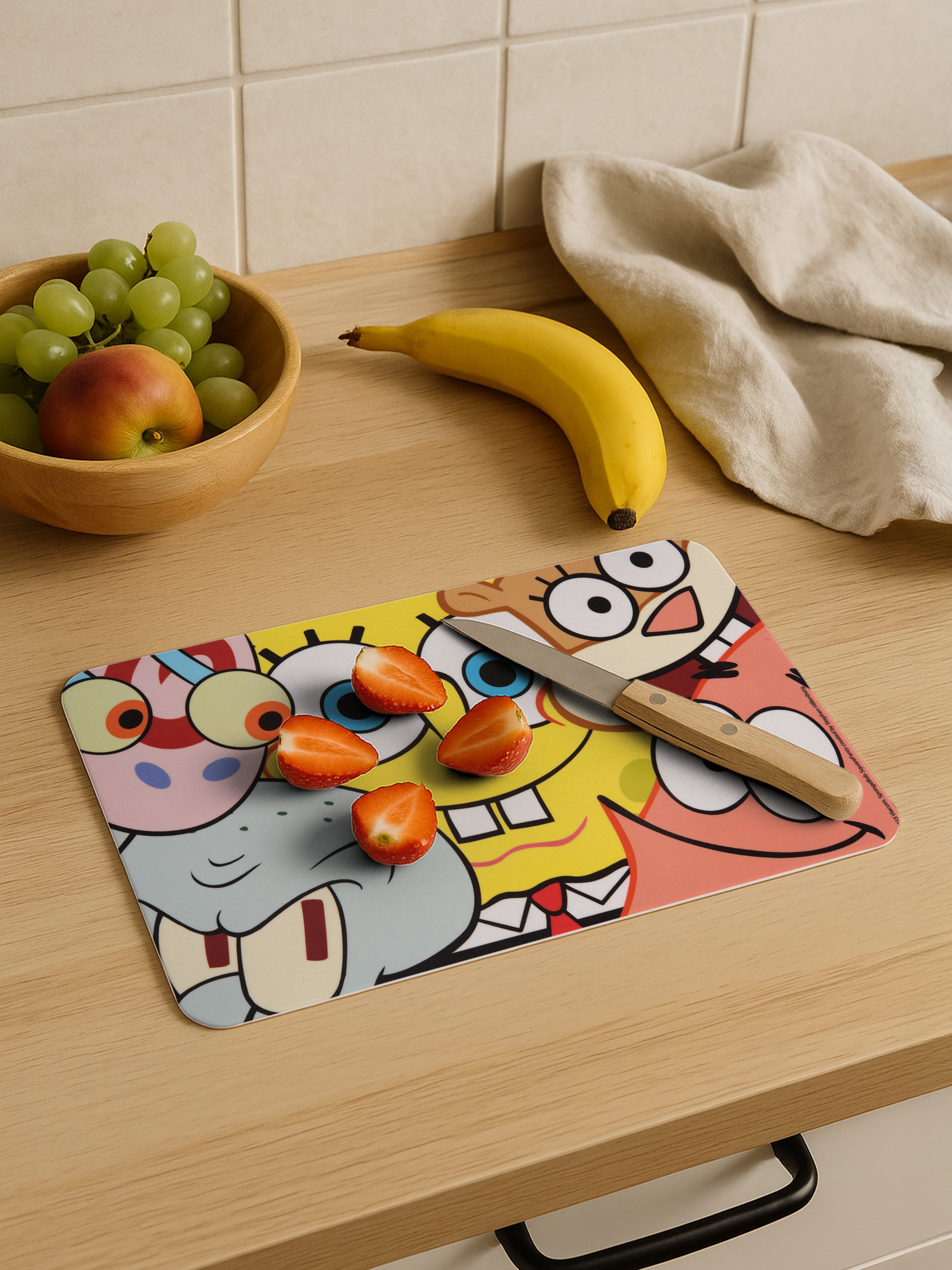 Ein Schneidebrett mit Cartoonfiguren, Erdbeeren, Banane und einem Messer liegt auf einer Holztheke mit Obst.